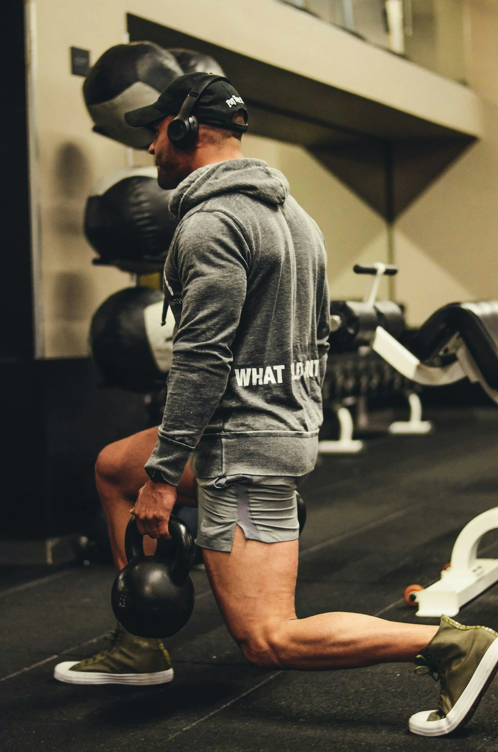 A man wearing a gray hoodie, gray shorts, and beige sneakers is kneeling on the ground and lifting a kettlebell in a gym. He is wearing a black cap and headphones, with a gym bag in the background.