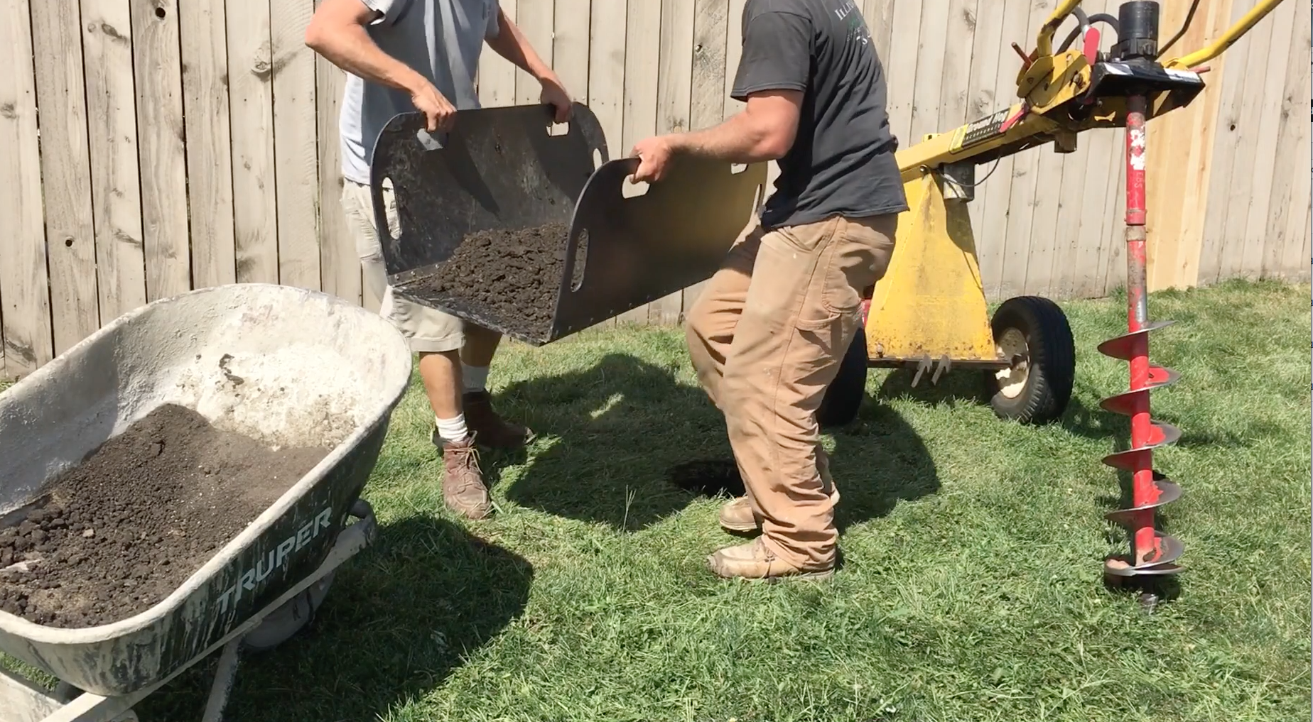 Two people filling a wheelbarrow with soil or dirt in a backyard, with a wood fence in the background and gardening equipment nearby.