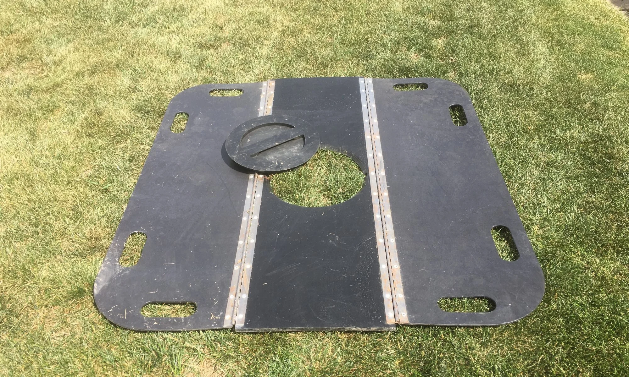 A black metal carburetor adapter plate with a circular opening and a smaller round, flat piece on top, placed on grass.