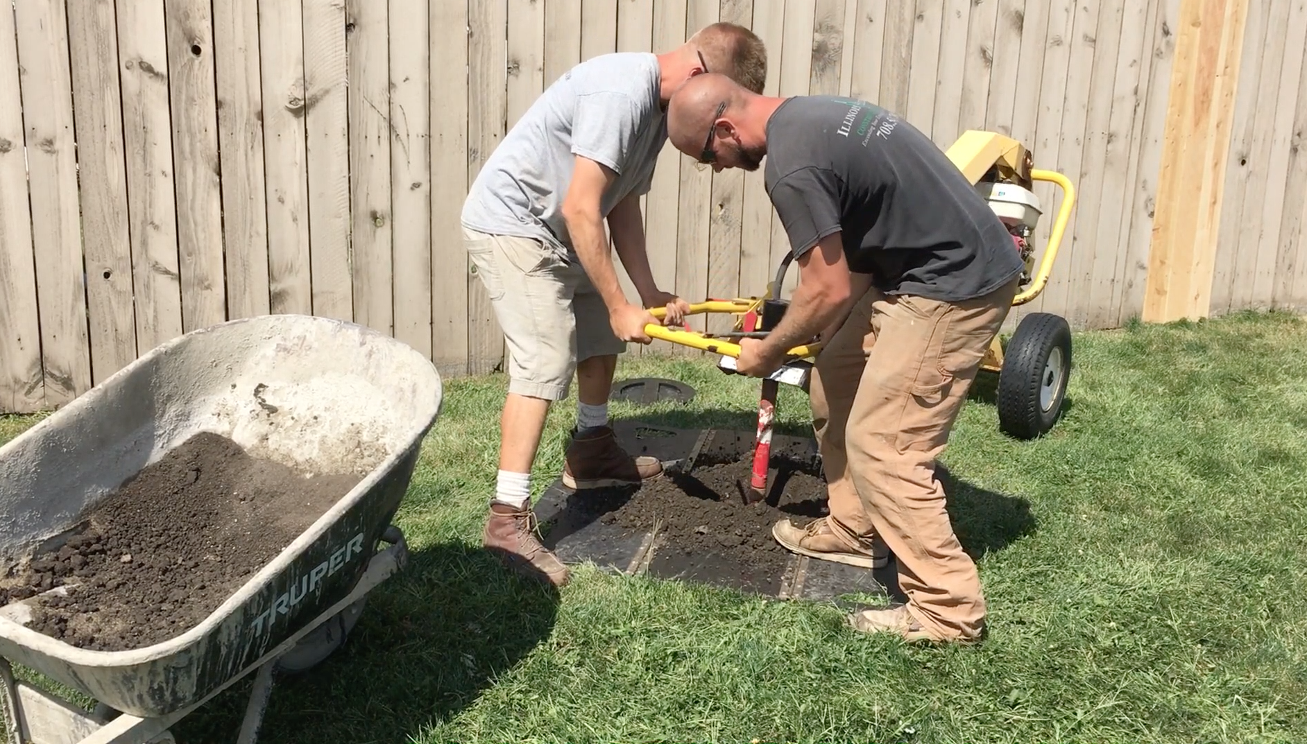 Two men planting a tree in a backyard using a post hole digger. One man is holding the handle and the other is operating the machine. There is a wheelbarrow filled with soil nearby and a wooden fence in the background.