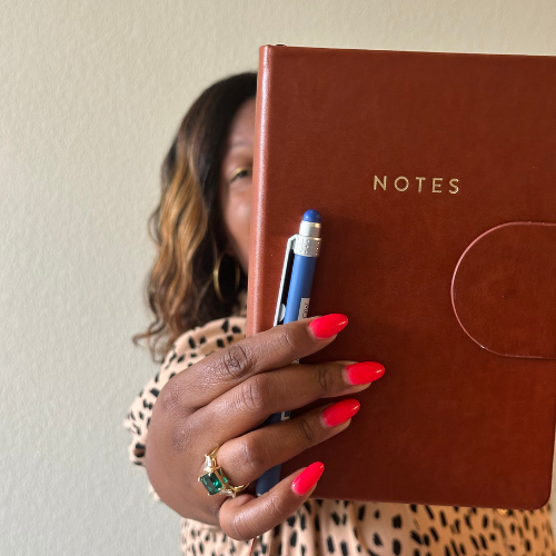 Person with dark skin and wavy hair holding a brown notebook titled Notes, with a pen and a ring with a green stone.