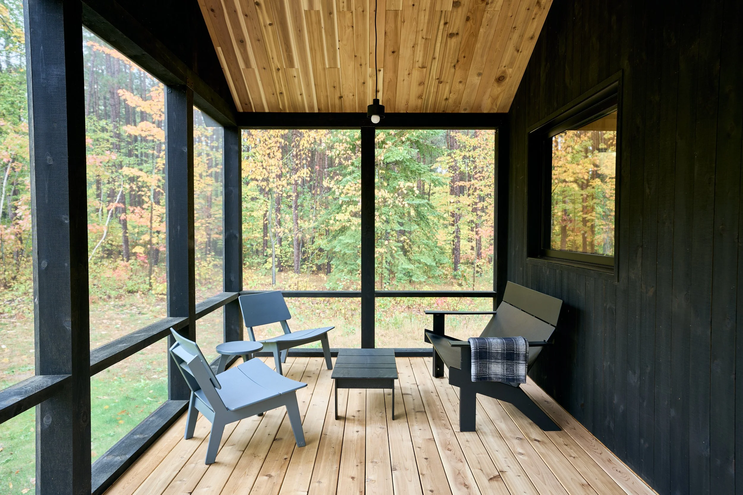 A screened porch with wooden flooring and ceiling, furnished with a bench, two chairs, and a small table, overlooking a forest with tall trees and green foliage.