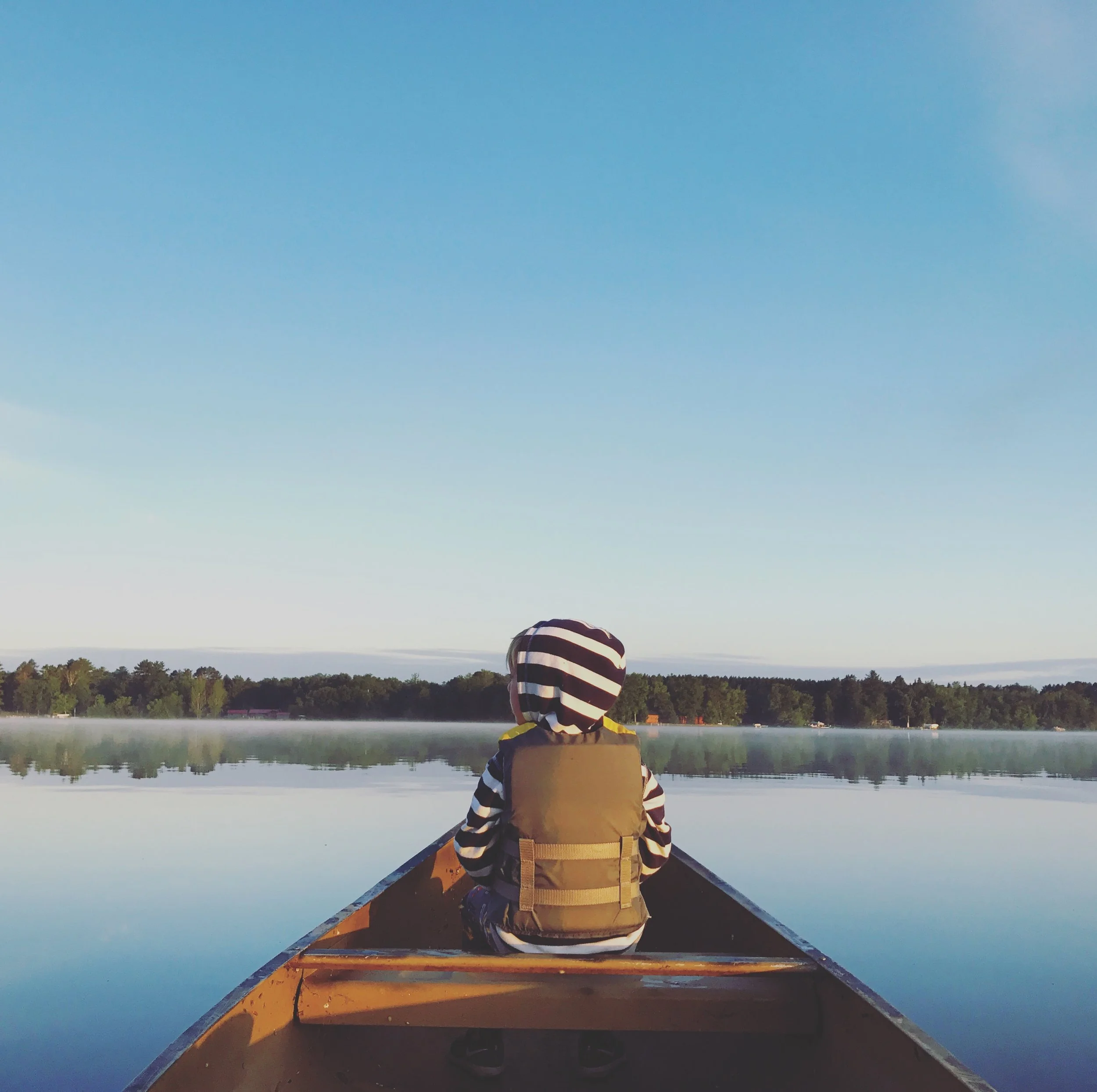 Person in striped hoodie and beige life jacket sits in a canoe on a calm lake, facing away, with forested shoreline in the distance and a clear blue sky above.