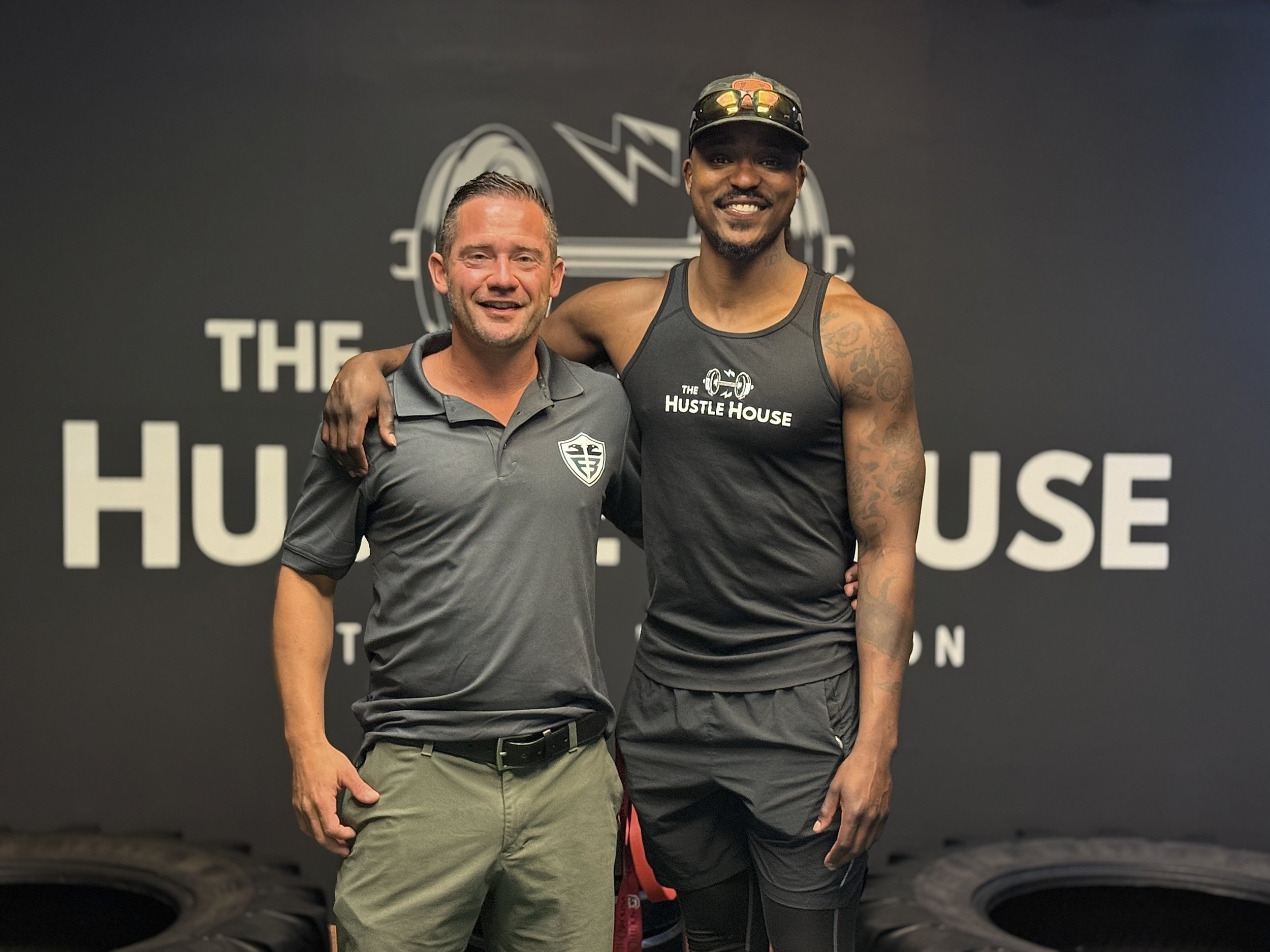 Two men standing together inside a gym, smiling at the camera. The man on the left is wearing a gray polo shirt with a logo and beige pants. The man on the right is wearing a black tank top with the words 'The Hustle House' and black shorts, with tattoos on his arms, sunglasses on his cap, and a beard.