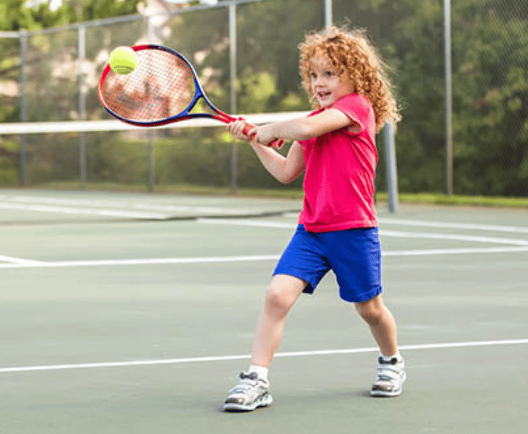 Young girl with curly hair playing tennis on an outdoor court, swinging a tennis racket at a yellow tennis ball.