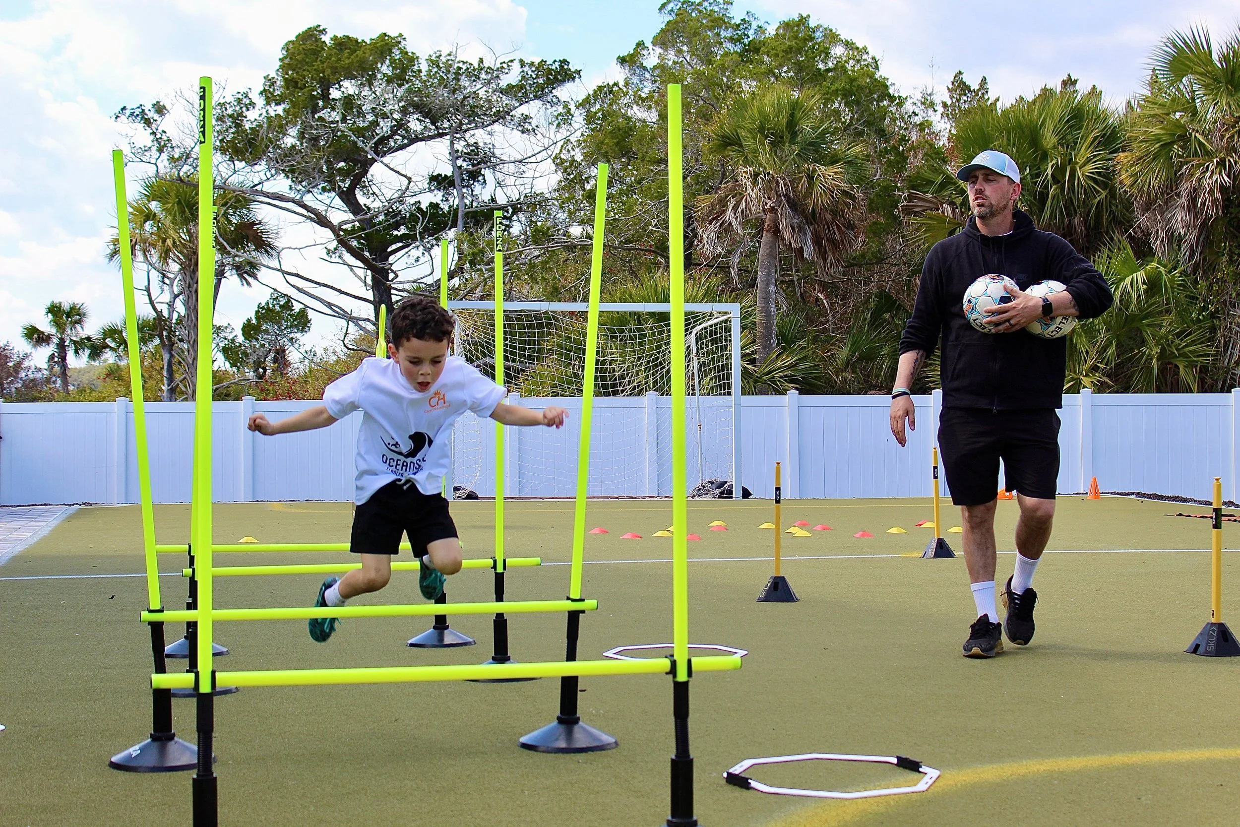 A young boy in a white shirt and black shorts is jumping over a hurdle during an outdoor sports training session. An adult coach, holding two soccer balls, is walking nearby on a field with agility equipment and cones, with a background of trees and a white fence.