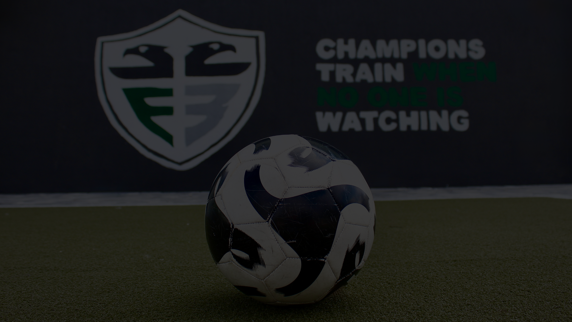 A soccer ball on an indoor field with a logo and a motivational quote on the wall behind it.