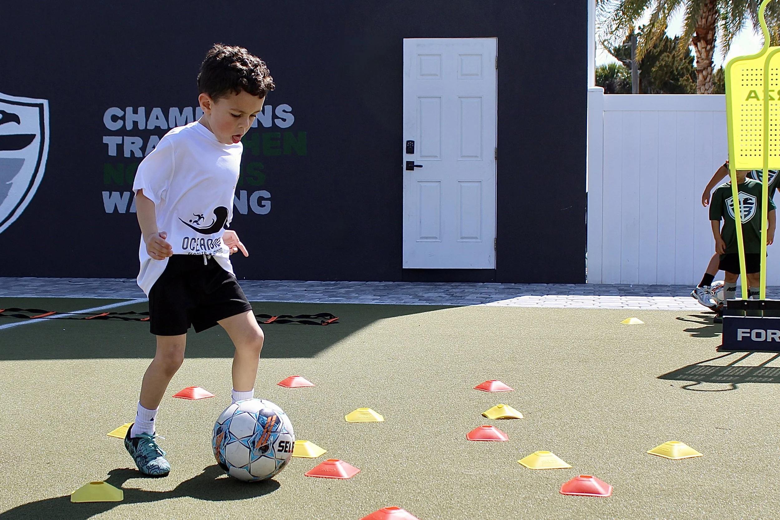 Young boy playing soccer on an outdoor field, dribbling a soccer ball among red and yellow training cones, with more children in the background near training equipment.
