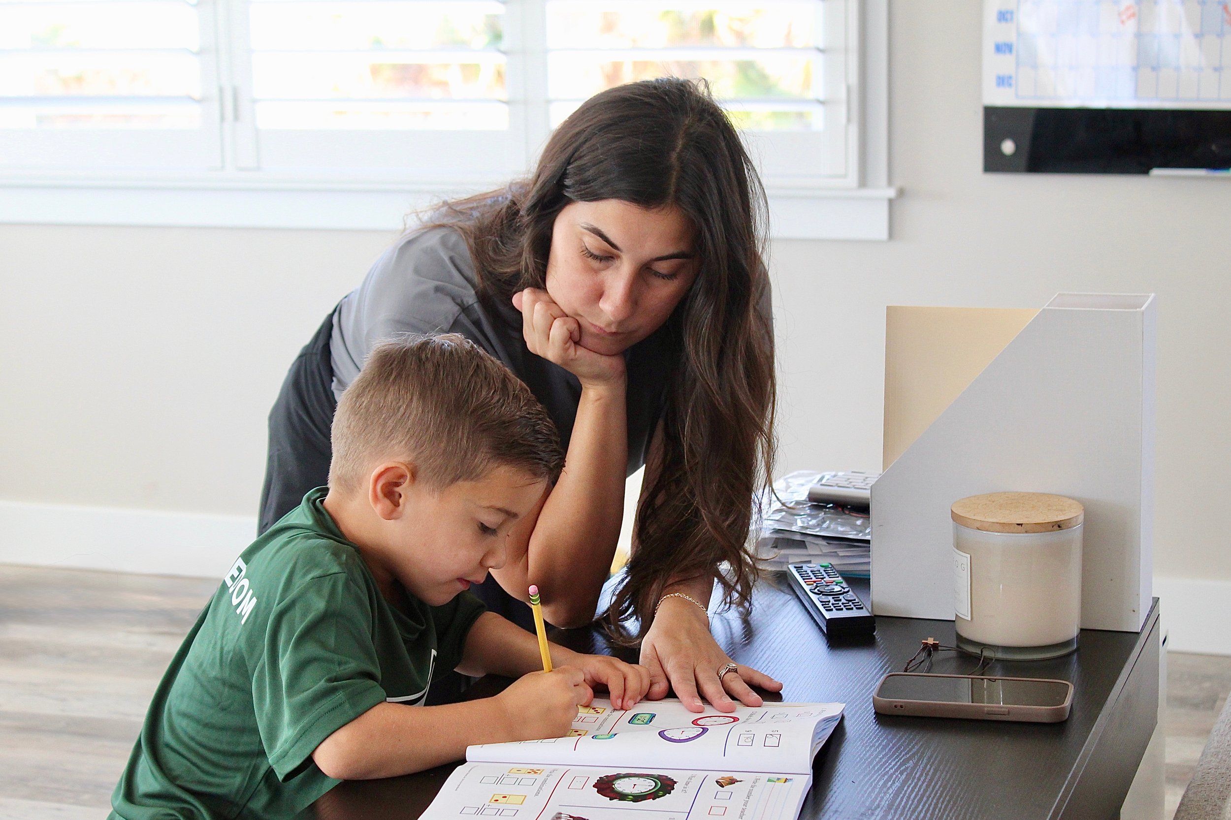 A woman helping a young boy with a homework assignment at a desk in a room with windows.