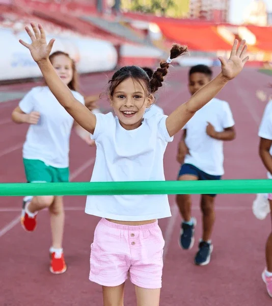 A young girl crossing the finish line in a race with her arms raised in celebration, surrounded by other children running behind her.