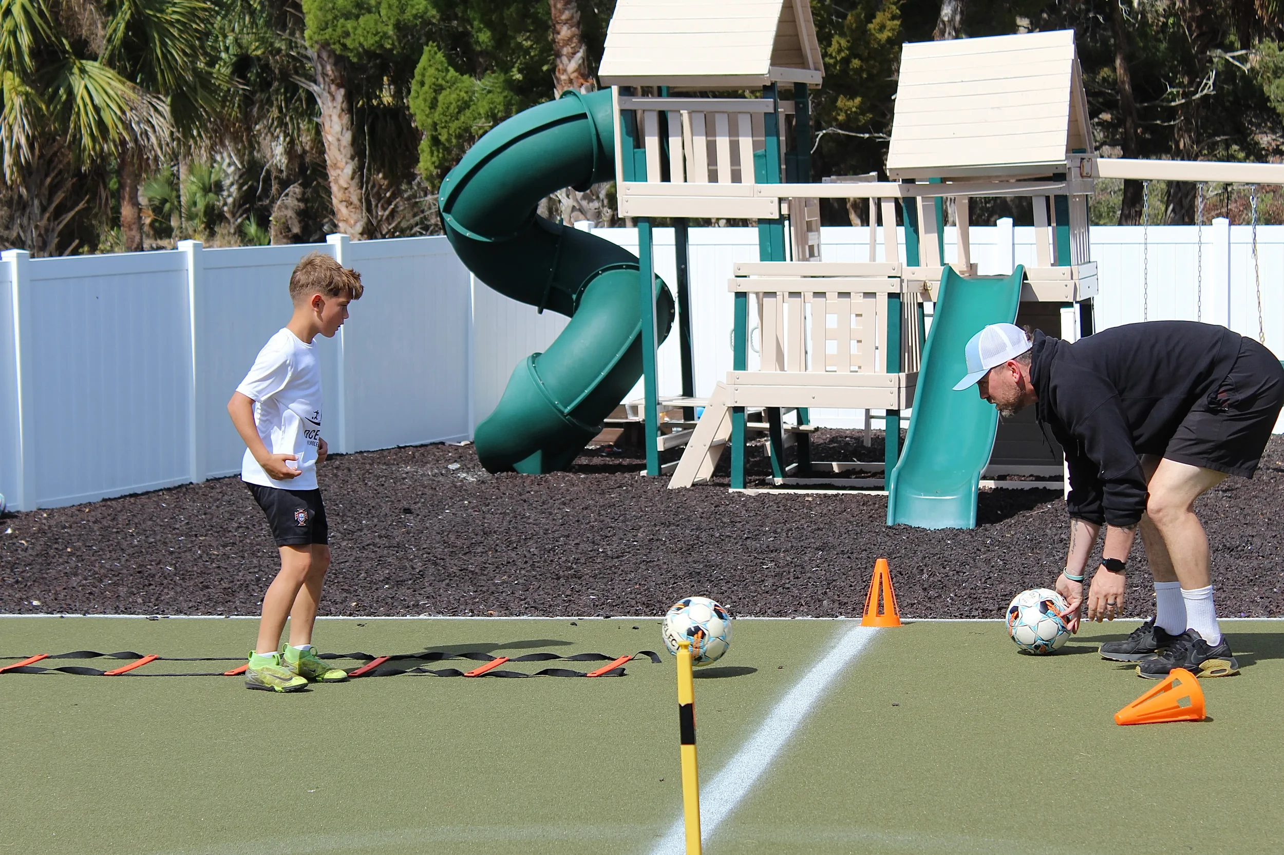 A young boy and an adult man preparing for a soccer drill on a green field with a playground and trees in the background. The boy is standing near agility ladders, and the man is crouched down adjusting a soccer ball. There are orange training cones and a pole with a soccer ball on top.