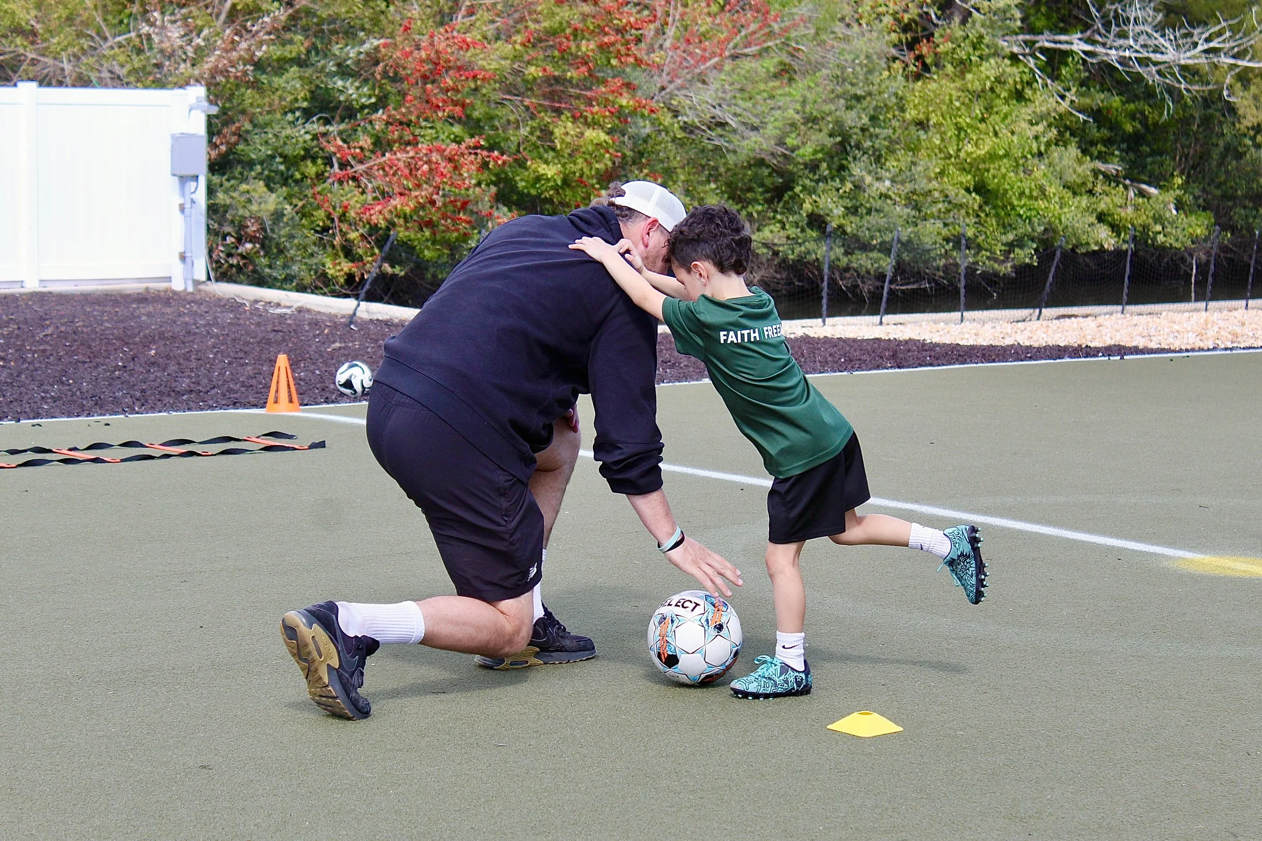 A man and a boy practicing soccer on a field. The man is bending down, and the boy is placing his hands on the man's shoulders and leaning forward. Soccer training equipment is scattered around, including a ball, cones, and an agility ladder, with trees in the background.