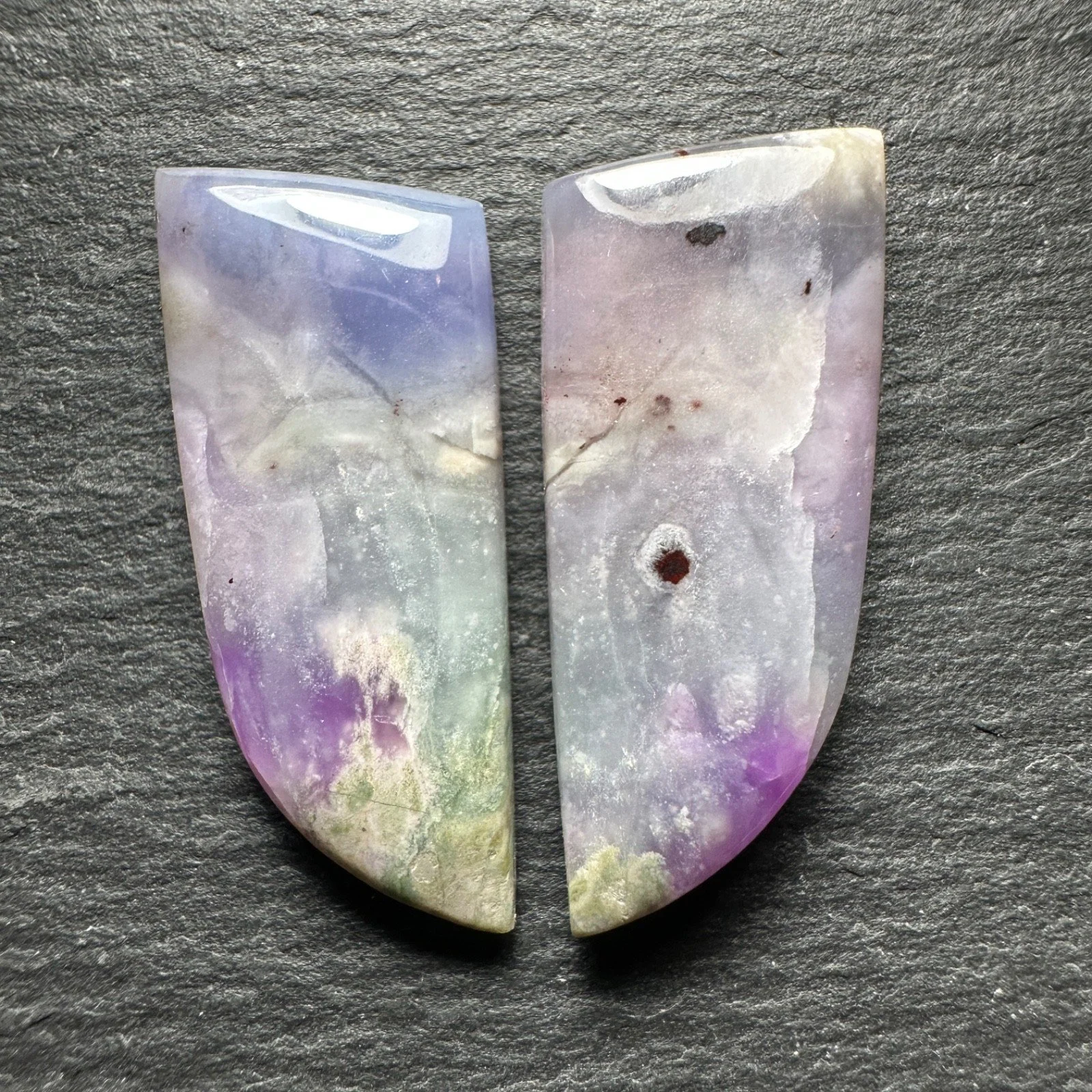 Two half pieces of a multicolored opal gemstone placed on a dark gray textured surface.