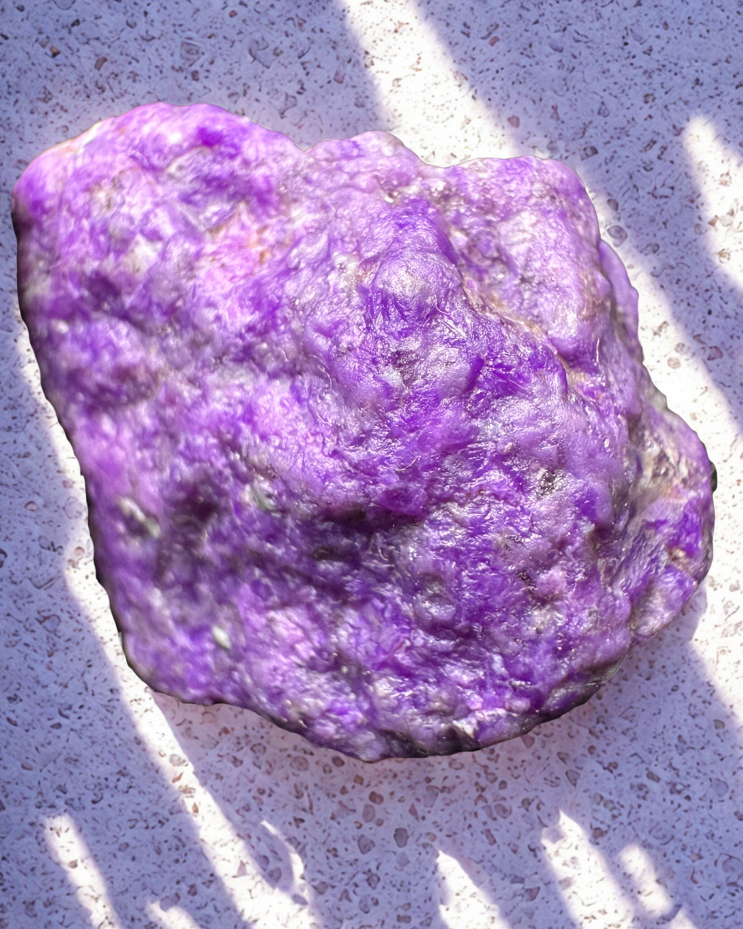 A large, irregularly shaped purple sugilite mineral or rock on a white textured surface with sunlight and shadows.