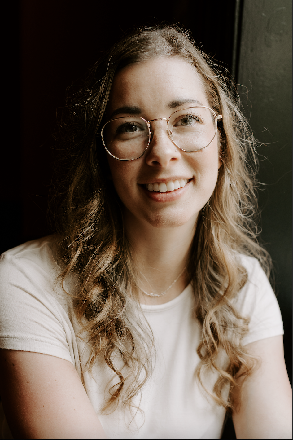 A smiling young woman with long wavy hair wearing glasses and a white t-shirt, sitting near a window.