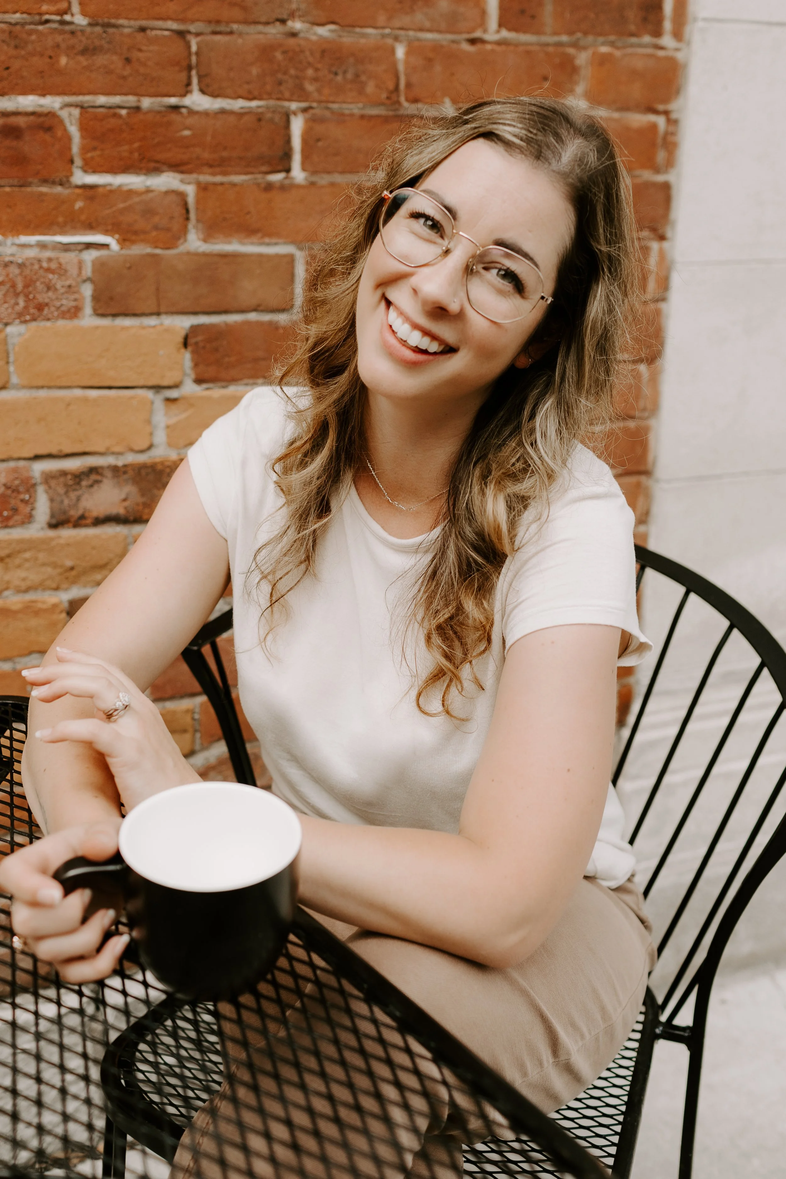 A young woman with glasses and wavy brown hair sitting at a black metal table, smiling, holding a black mug, against a brick wall background.