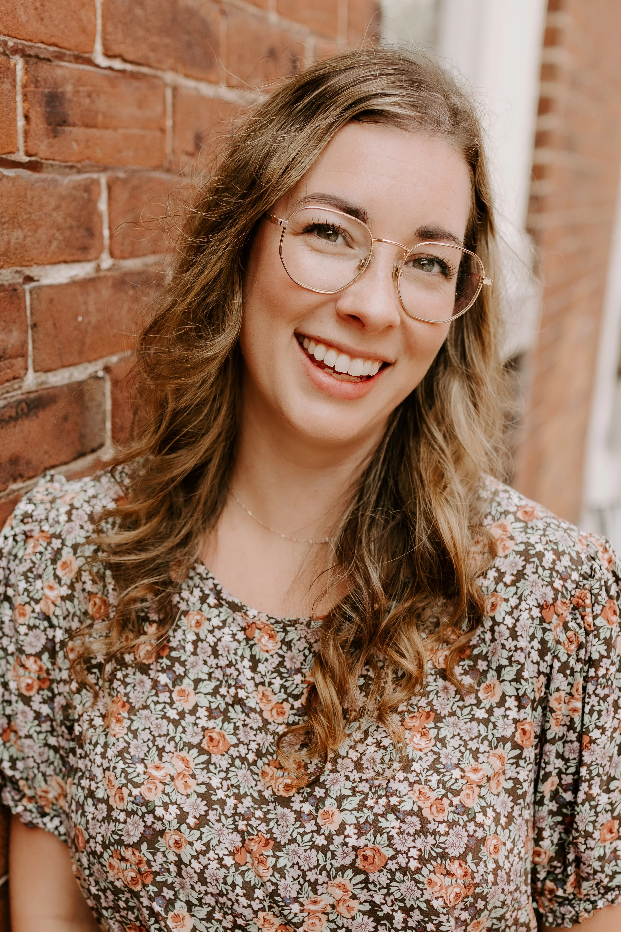A young woman with long curly hair and glasses, smiling, standing against a brick wall, wearing a floral patterned shirt.