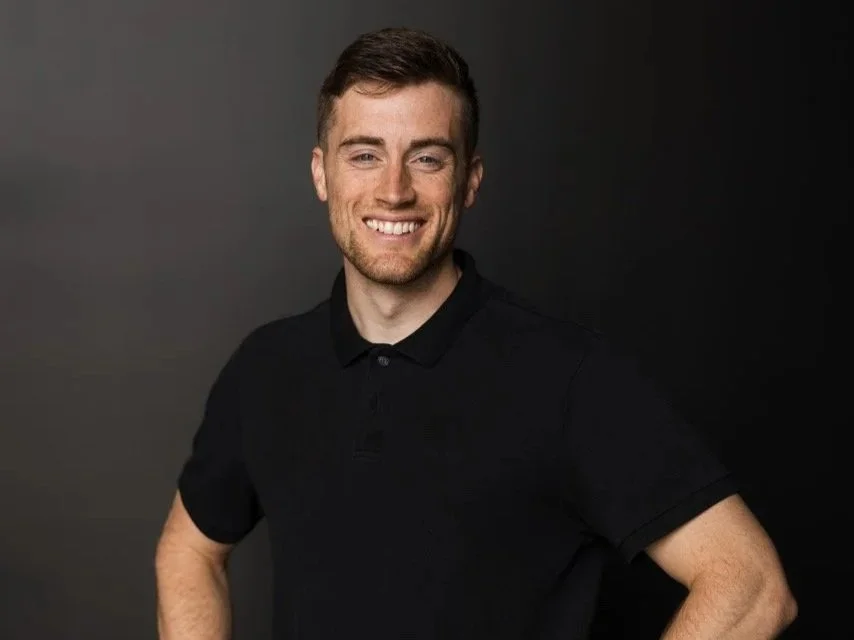 A young man with brown hair and a beard smiling and crossing his arms, wearing a black t-shirt, against a plain white background.