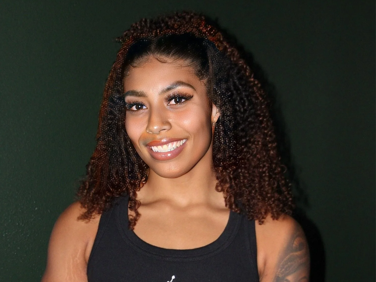 A smiling woman with curly hair and brown skin, wearing a black sleeveless top, standing against a dark green background.