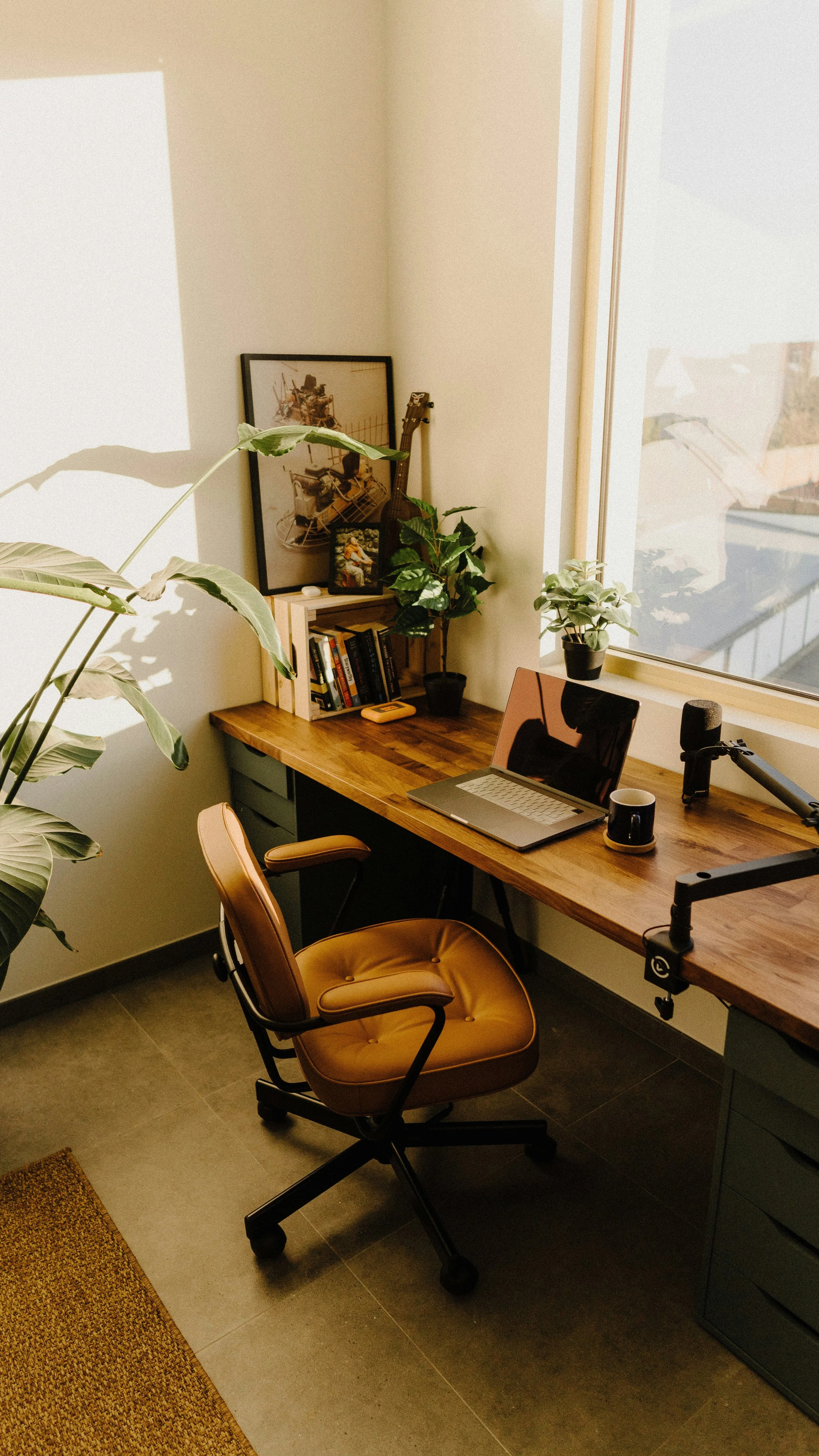 A home office with a wooden desk, a tan office chair, a laptop, a cup, a microphone, and plants near a window.