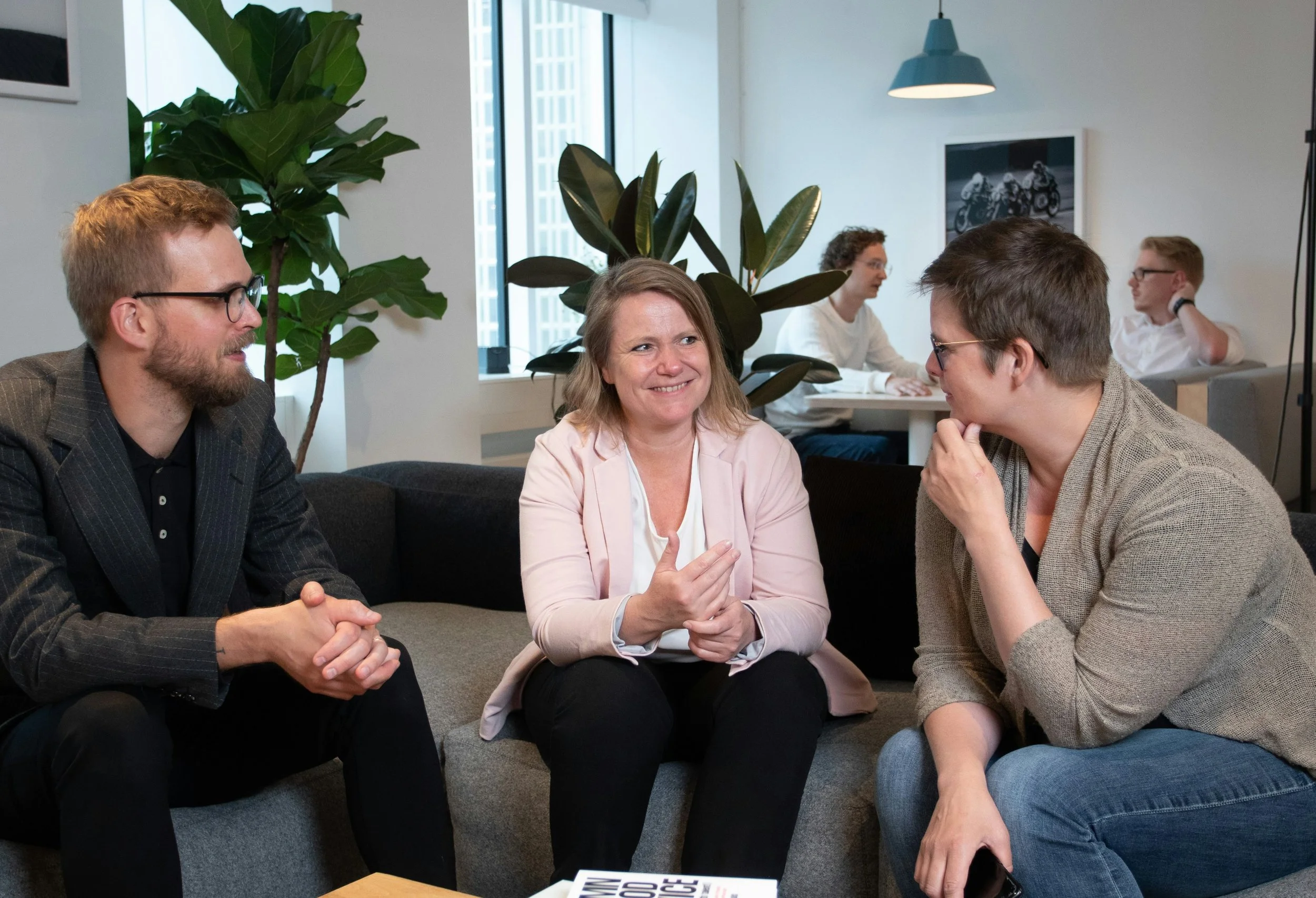 Group of four people sitting on a gray couch in an office, engaged in conversation. The woman in the center is smiling, wearing a light pink blazer, with two people on either side of her and two others working at a table in the background.