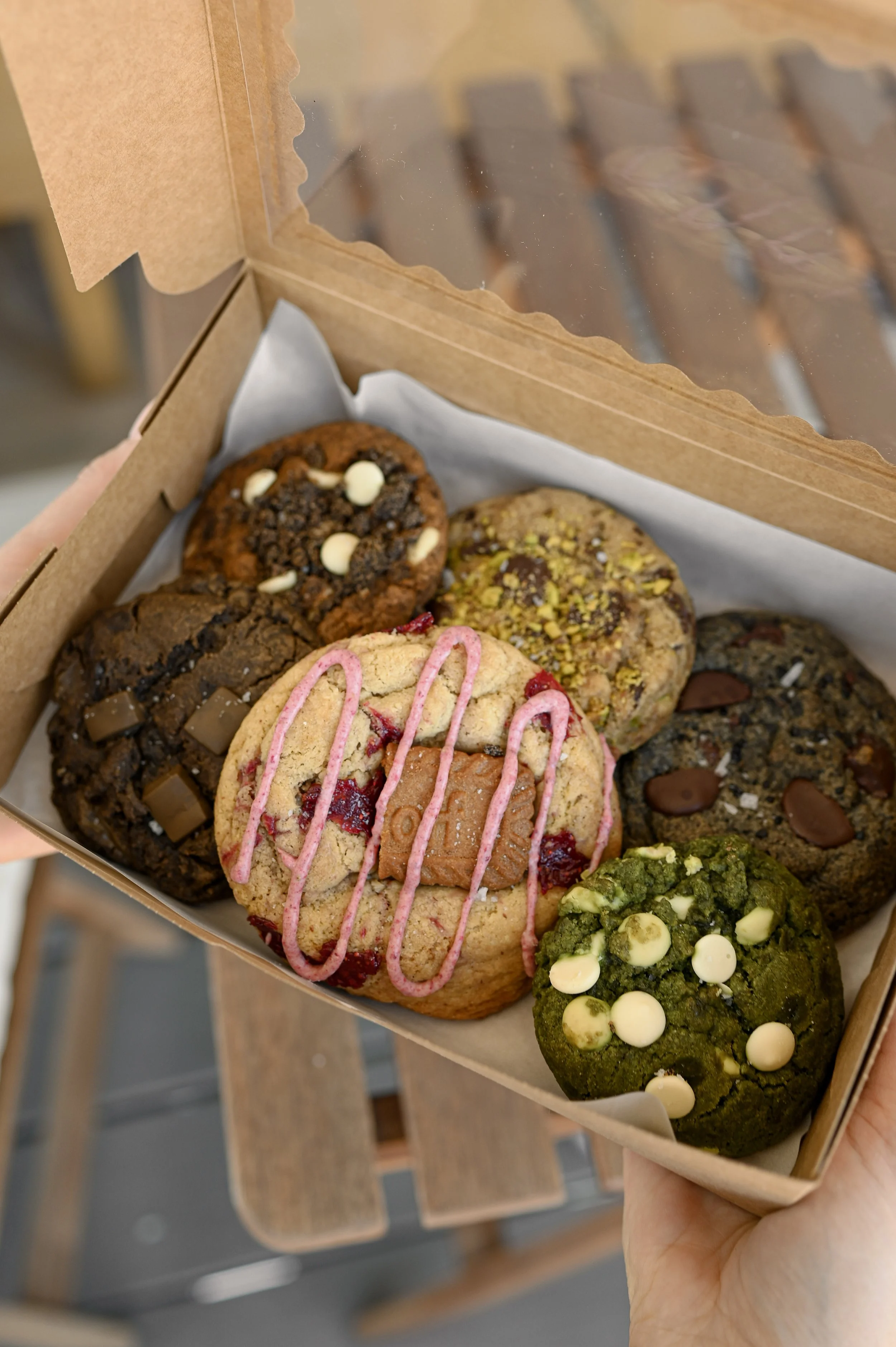 Hands holding a bakery box filled with an assortment of six Little Wins Bakehouse cookies lined with parchment paper.