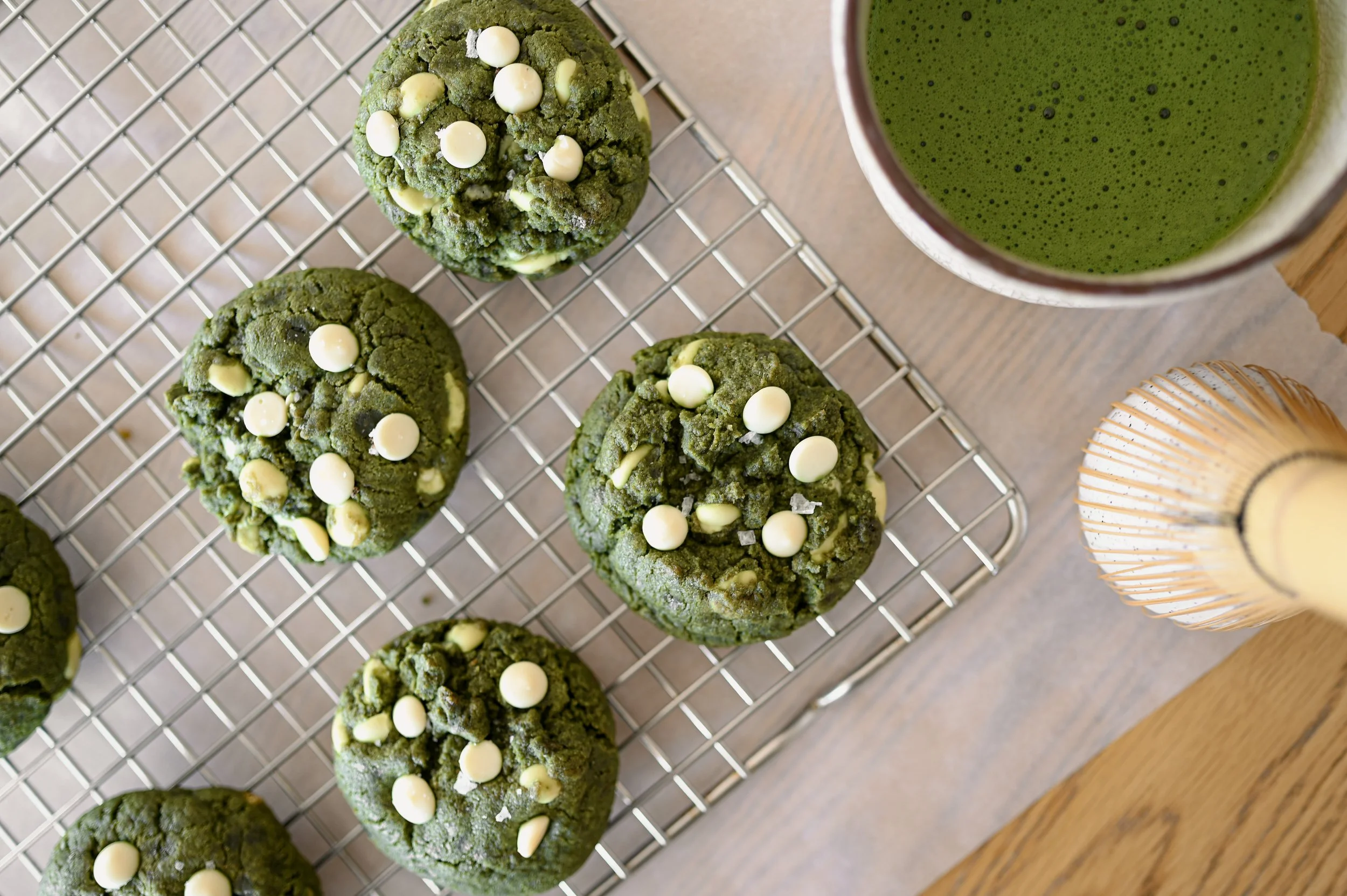 Matcha white chocolate cookies on a cooling rack alongside a bowl of freshly whisked matcha and a bamboo matcha whisk.