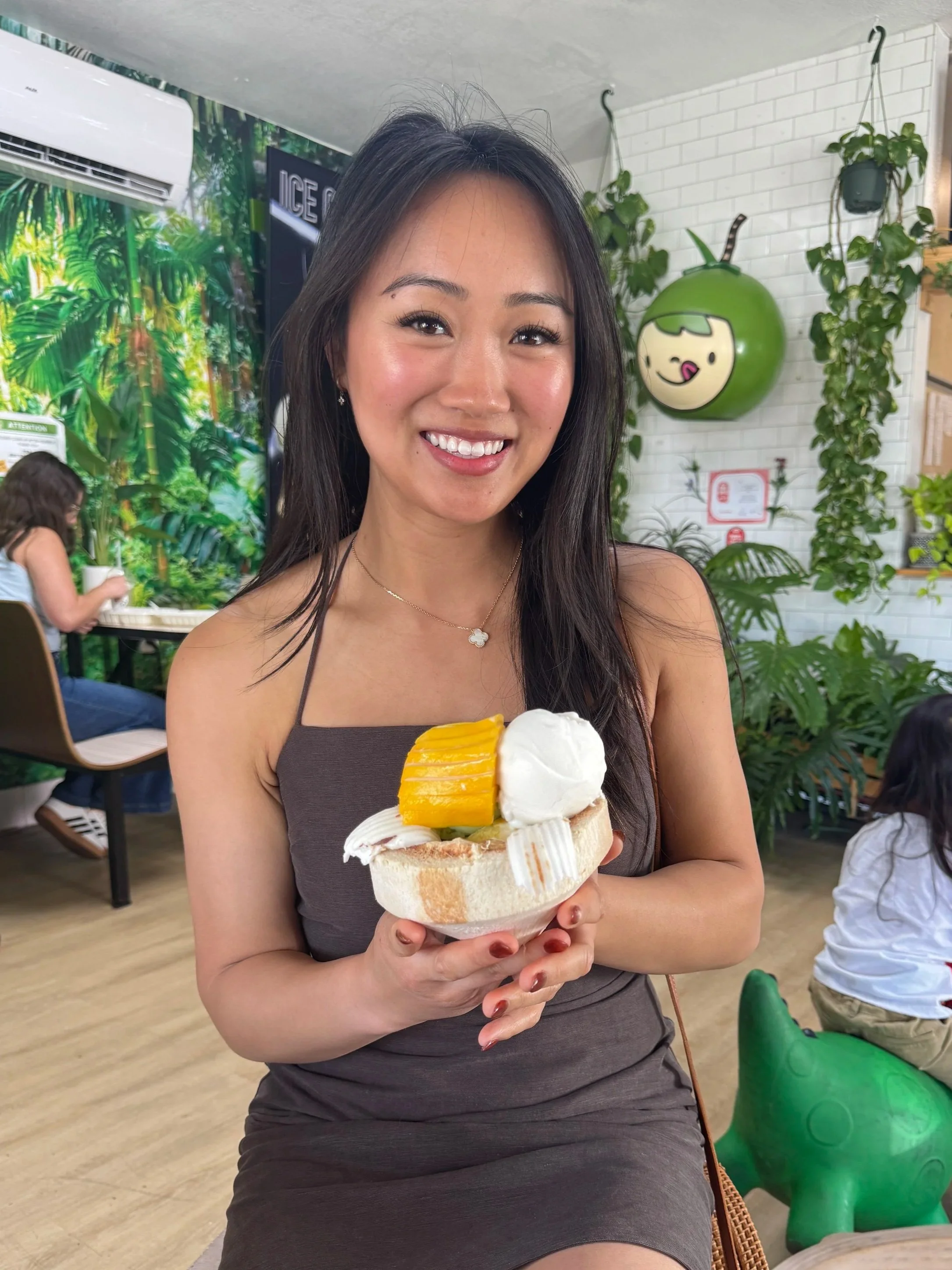 Win, the baker behind Little Wins Bakehouse, holding a coconut dessert inside a bright dessert café with plants and decorated walls.