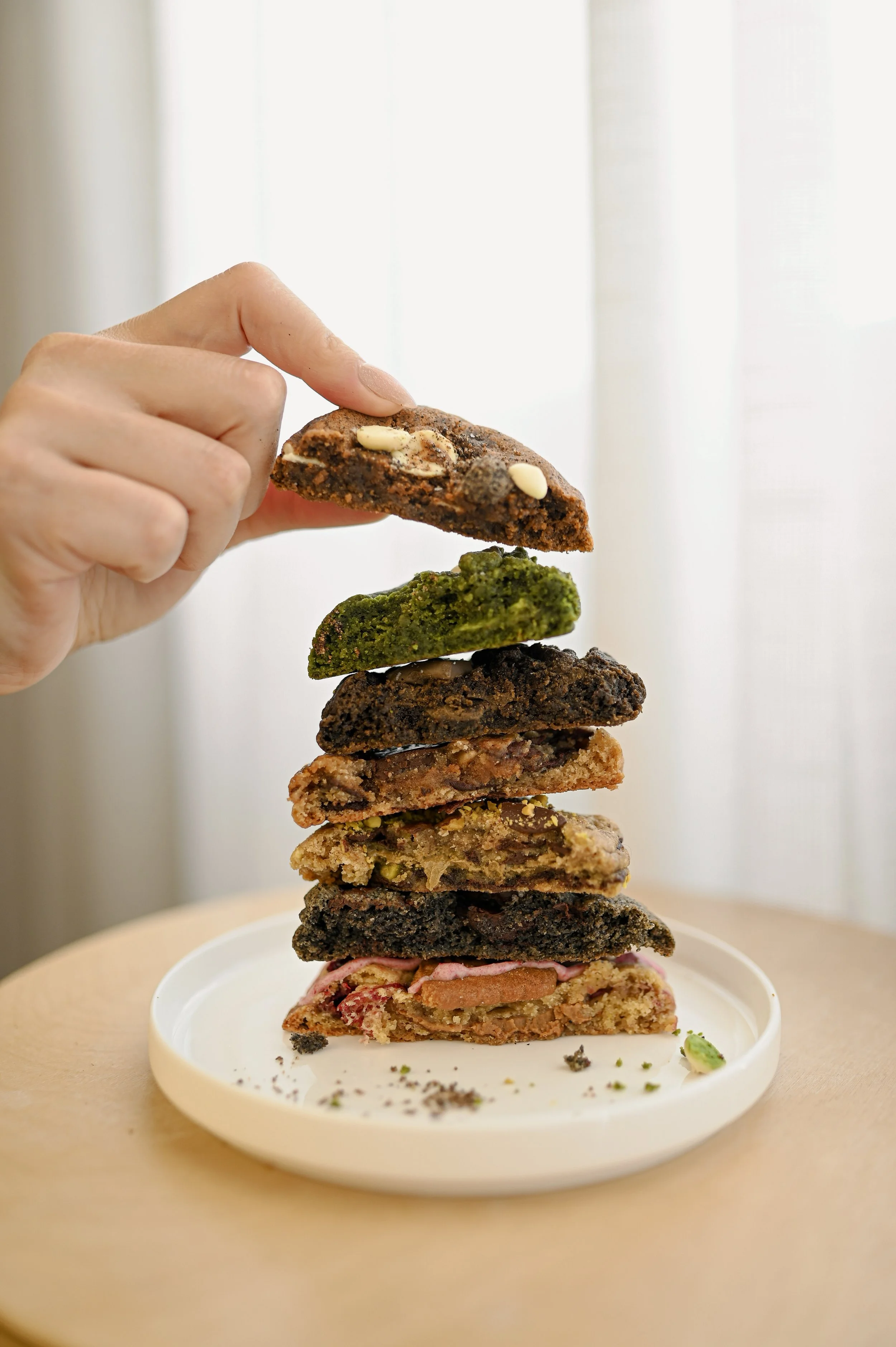 Hand stacking assorted cookie halves on a white plate, showing the soft centers and fillings of various Little Wins Bakehouse cookies.