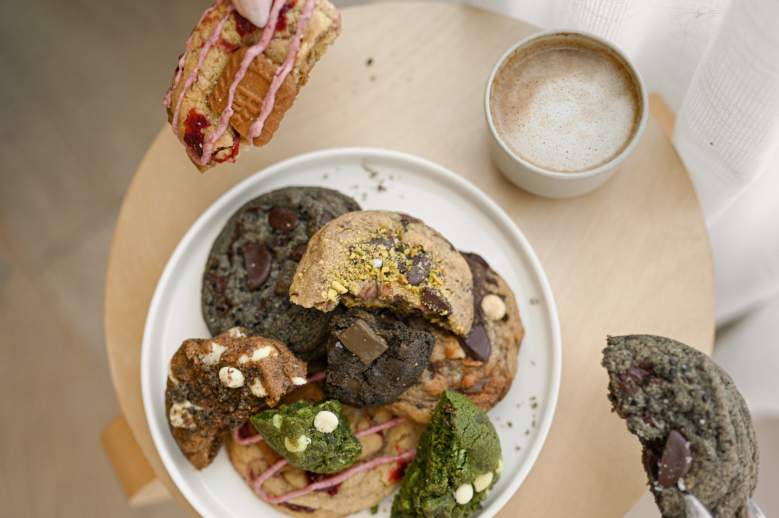 Assorted Little Wins Bakehouse cookies arranged on a white plate beside a latte on a wooden table.