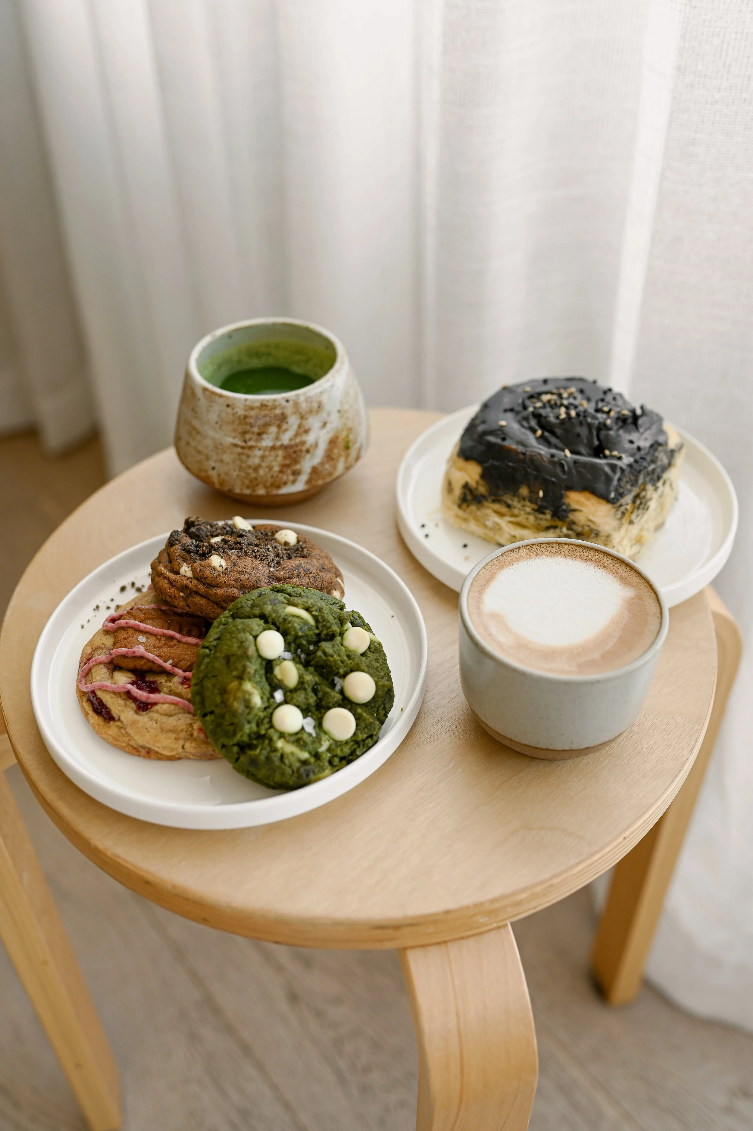 Assortment of Little Wins Bakehouse treats including cookies, a black sesame cinnamon roll, matcha, and coffee on a wooden table in front of white curtains.