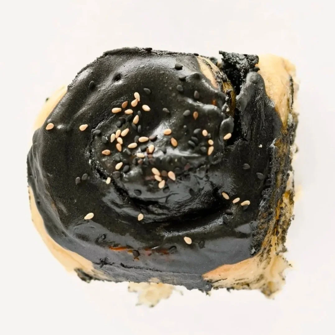 Black sesame cinnamon roll topped with black sesame buttercream and black and white sesame seeds.