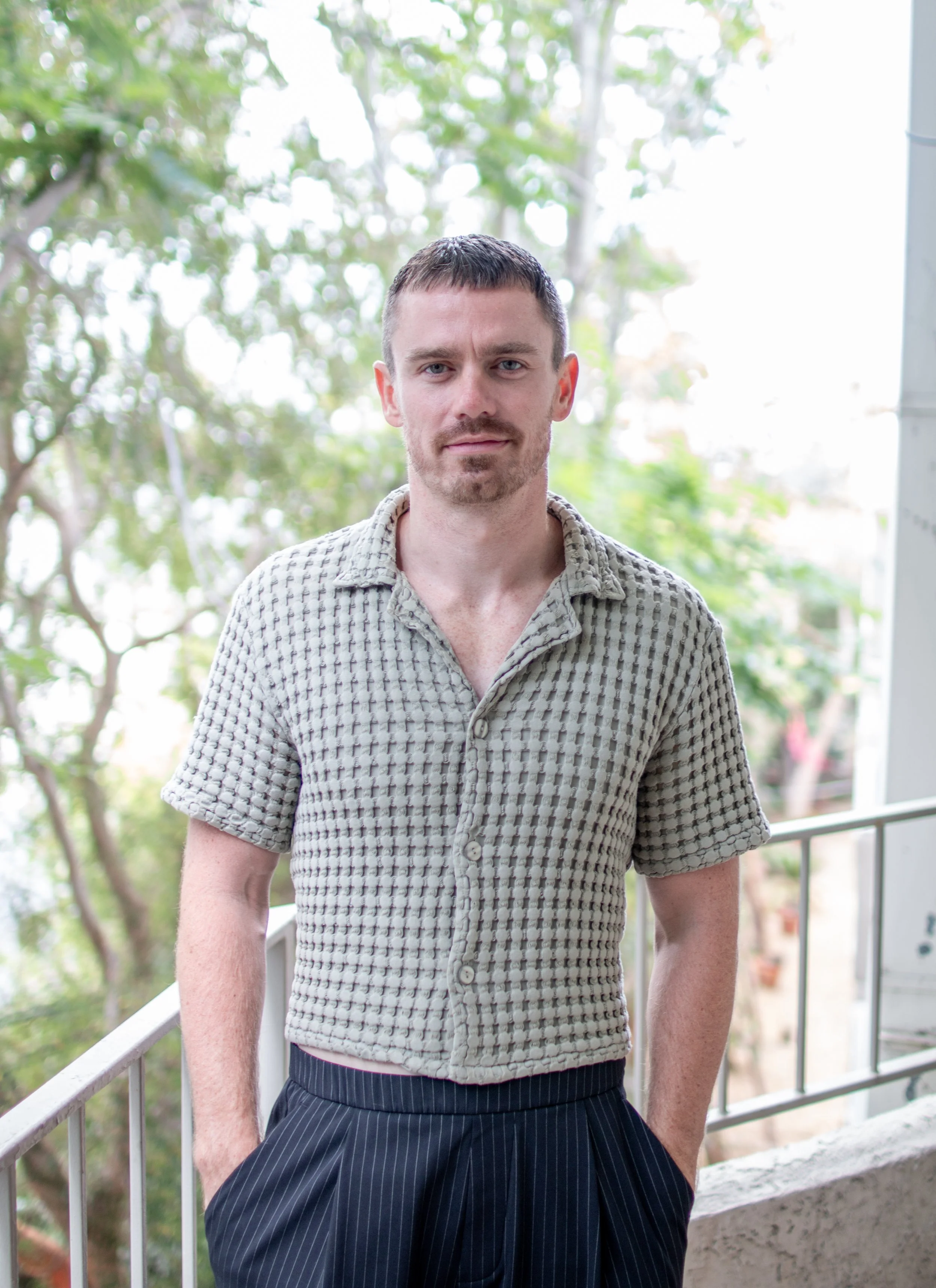 A man with short dark hair and a light beard stands outdoors on a balcony with a background of green trees. He wears a beige short-sleeved shirt with a textured pattern and dark pinstriped pants. His hands are in his pockets, and he is looking at the camera.