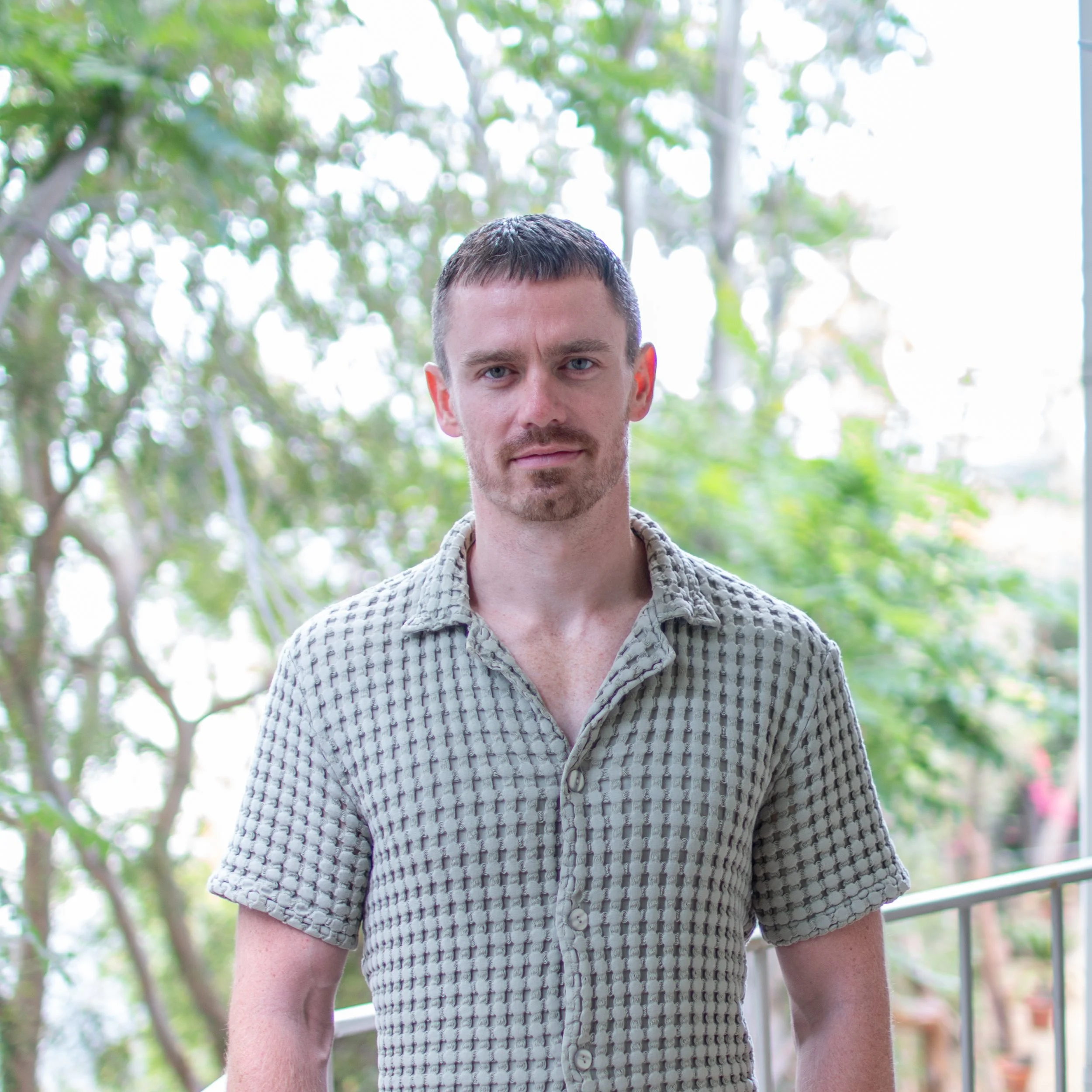 Close-up of a young man with short dark hair and blue eyes, wearing a textured light-colored short-sleeve shirt, standing outdoors with green trees and sunlight in the background.