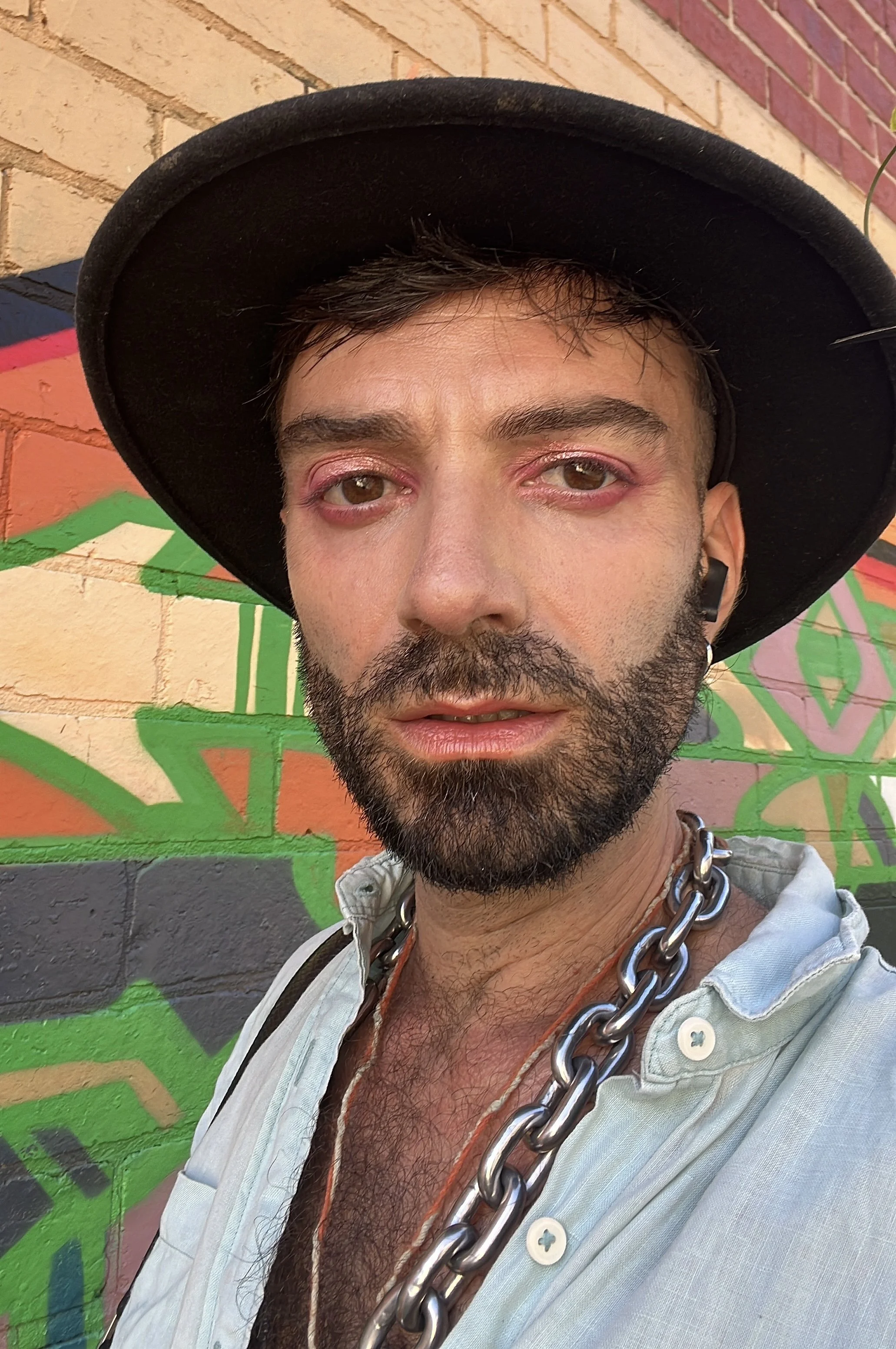 Profile picture of Ludo with a beard, colourful make up, a black hat, a chunky chain necklace, light-colored shirt, standing in front of a colorful graffiti wall.