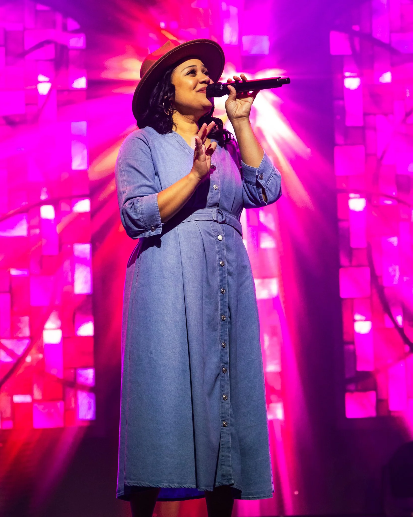 A woman singing on stage with colorful pink and purple lights in the background, wearing a denim dress and a wide-brimmed hat.