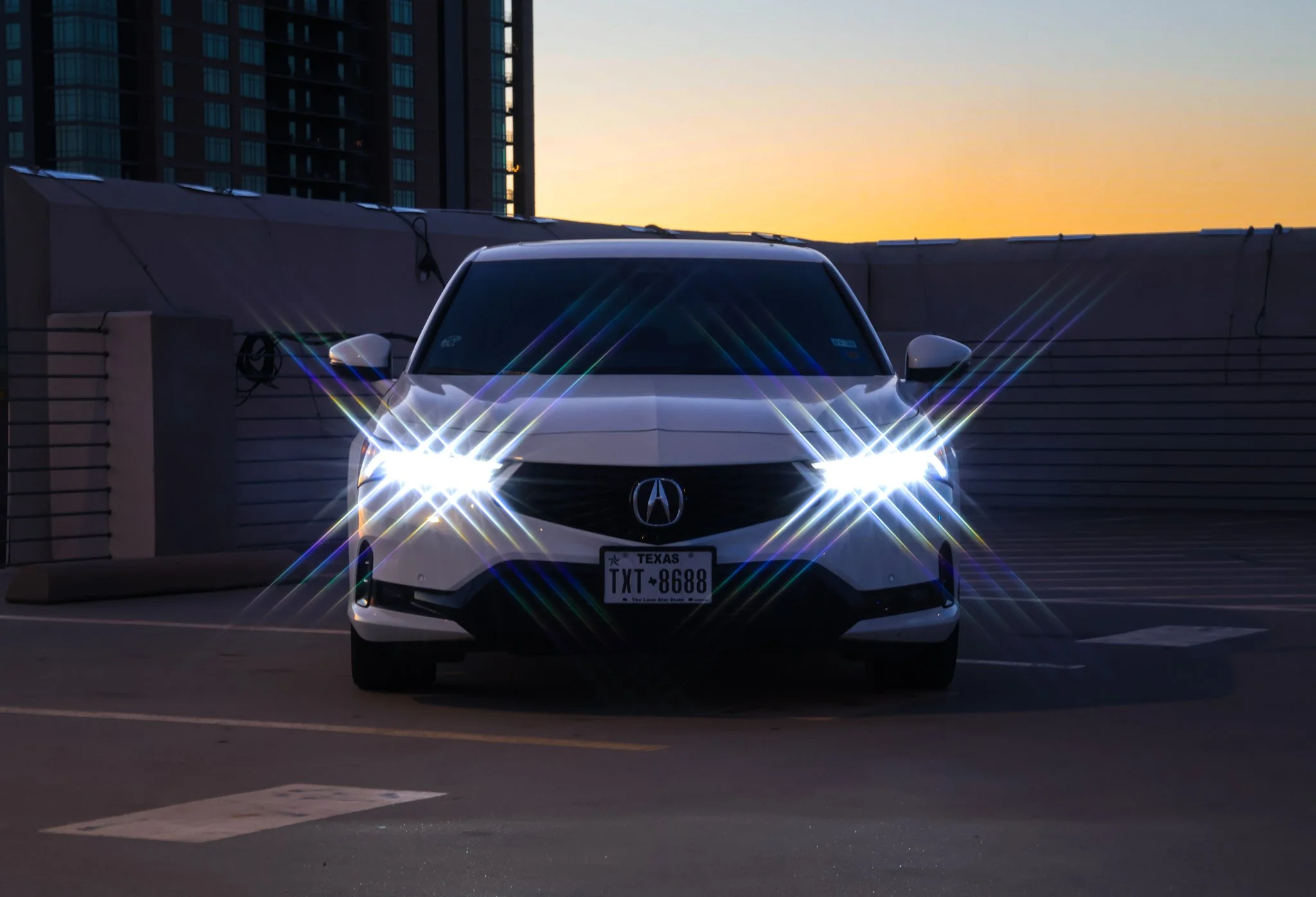 White Acura car with bright headlights parked on a rooftop parking lot at sunset, with a city skyline in the background.