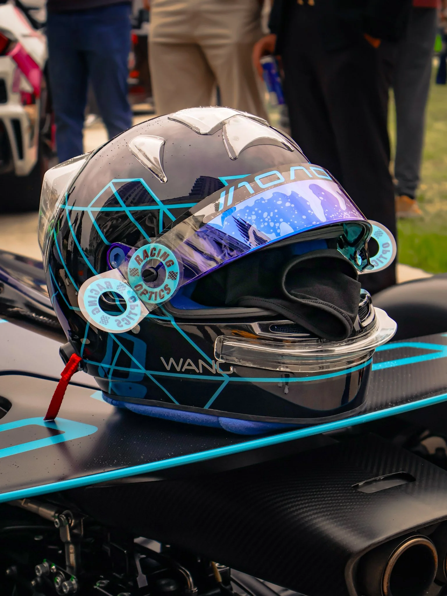 A racing helmet with a blue and black geometric design, placed on a racing motorcycle, surrounded by people at an outdoor event.