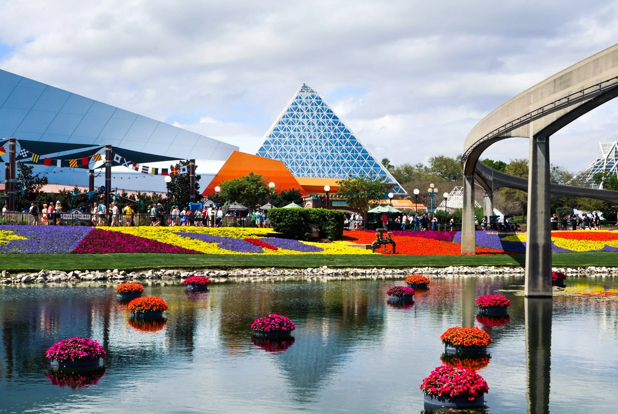 People walking in front of colorful geometric buildings at an amusement park with a flower garden and pond in the foreground.