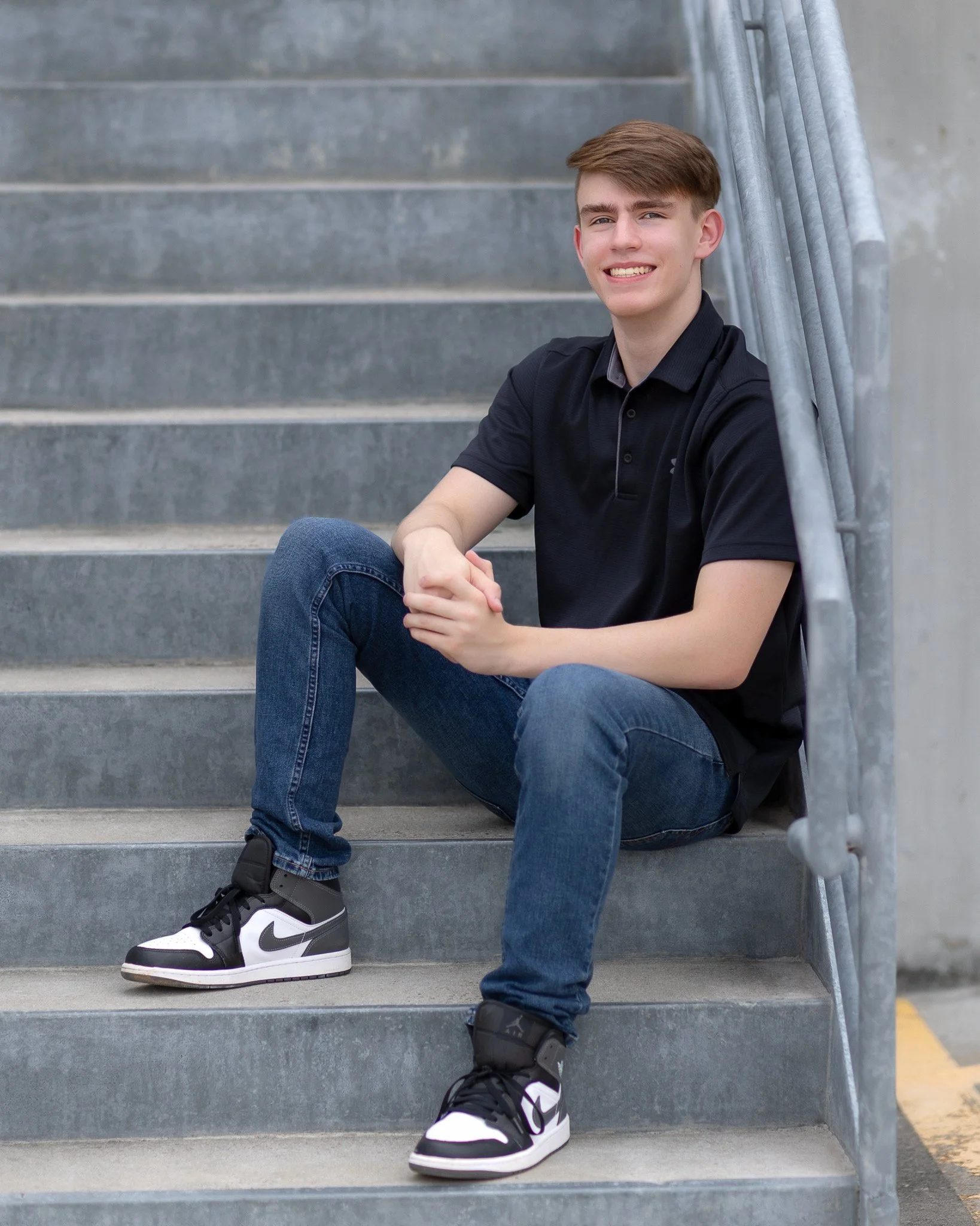 A young man sitting on outdoor concrete stairs, smiling, wearing a black polo shirt, blue jeans, and black-and-white Nike sneakers, with a metal railing on his right side.