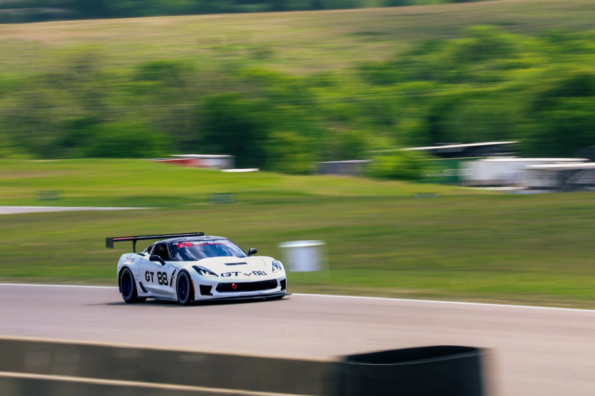 A white race car with black accents and the number GT 88 racing on a track, speeding past with a blurred background of green grassy hills and trees.