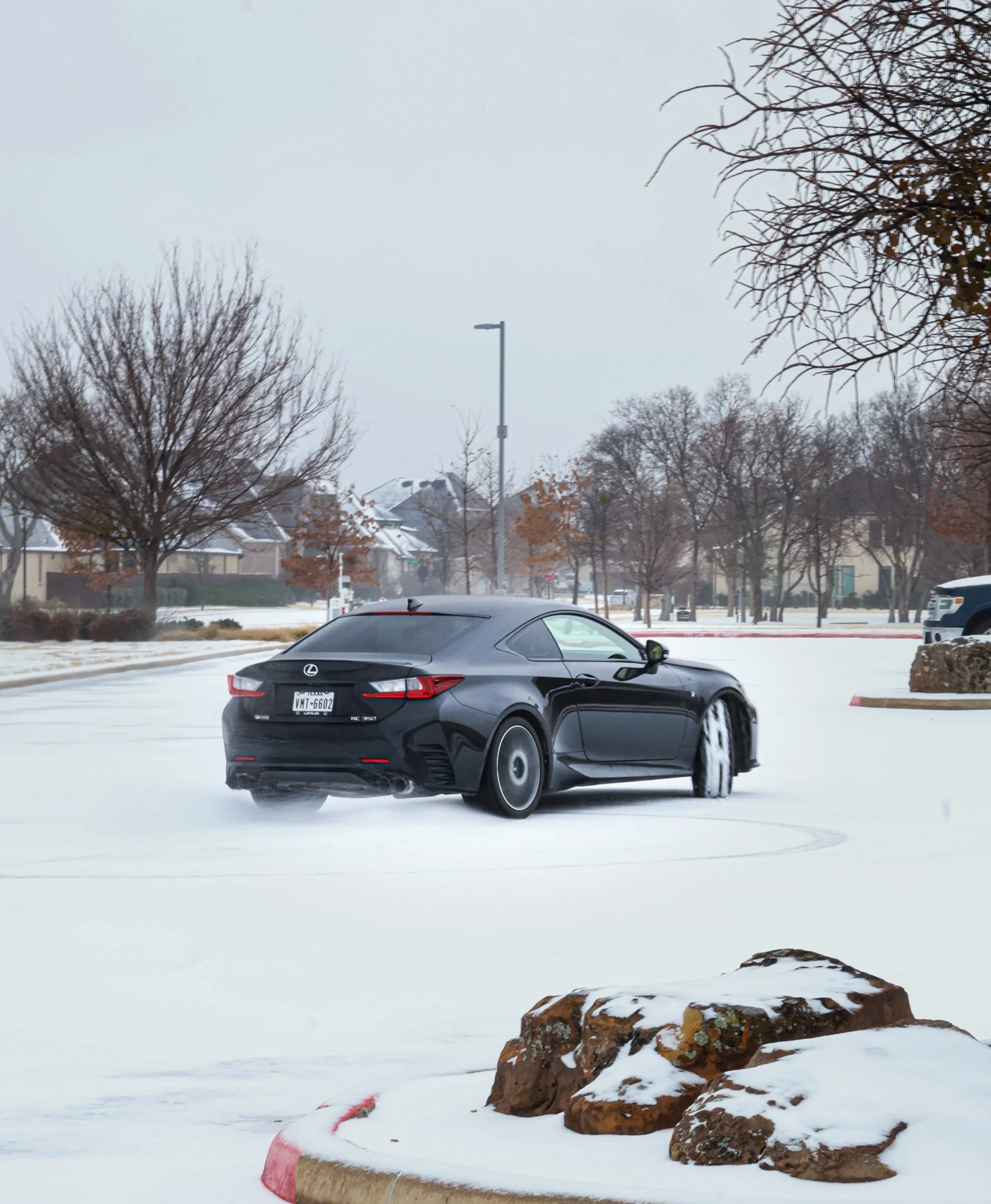 A black Lexus car on a snowy parking lot during winter day with trees and houses in the background.