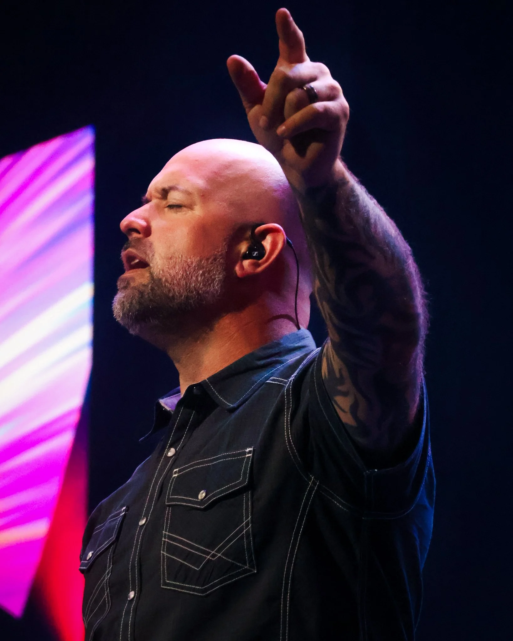 A man singing or speaking with his eyes closed, wearing a black button-up shirt, with a tattooed arm raised, and an in-ear monitor in his ear, on stage with colorful lights in the background.