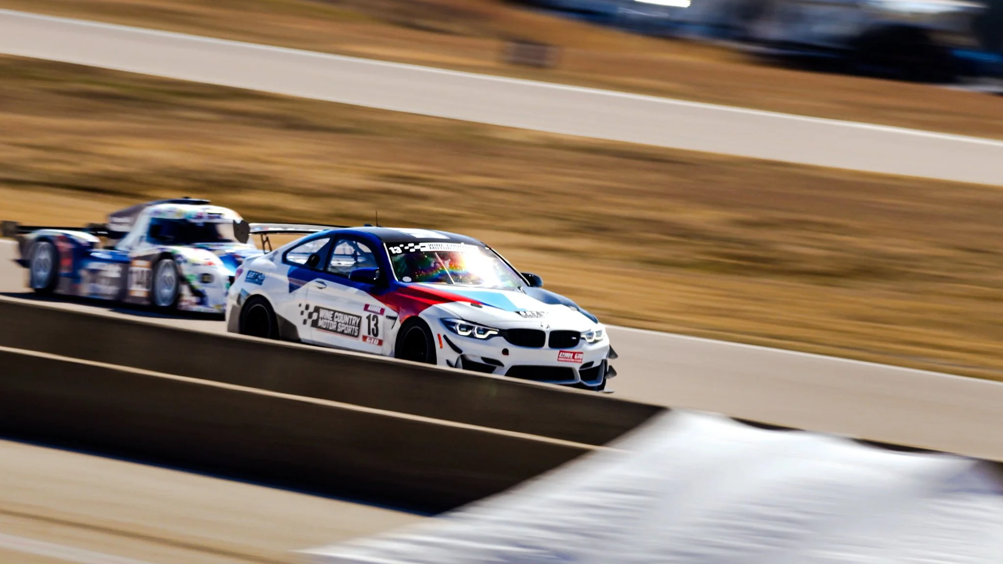 A white race car with red and blue accents, number 13, on a race track, leading a blue and white race car in the background, with a blurred background indicating high speed.