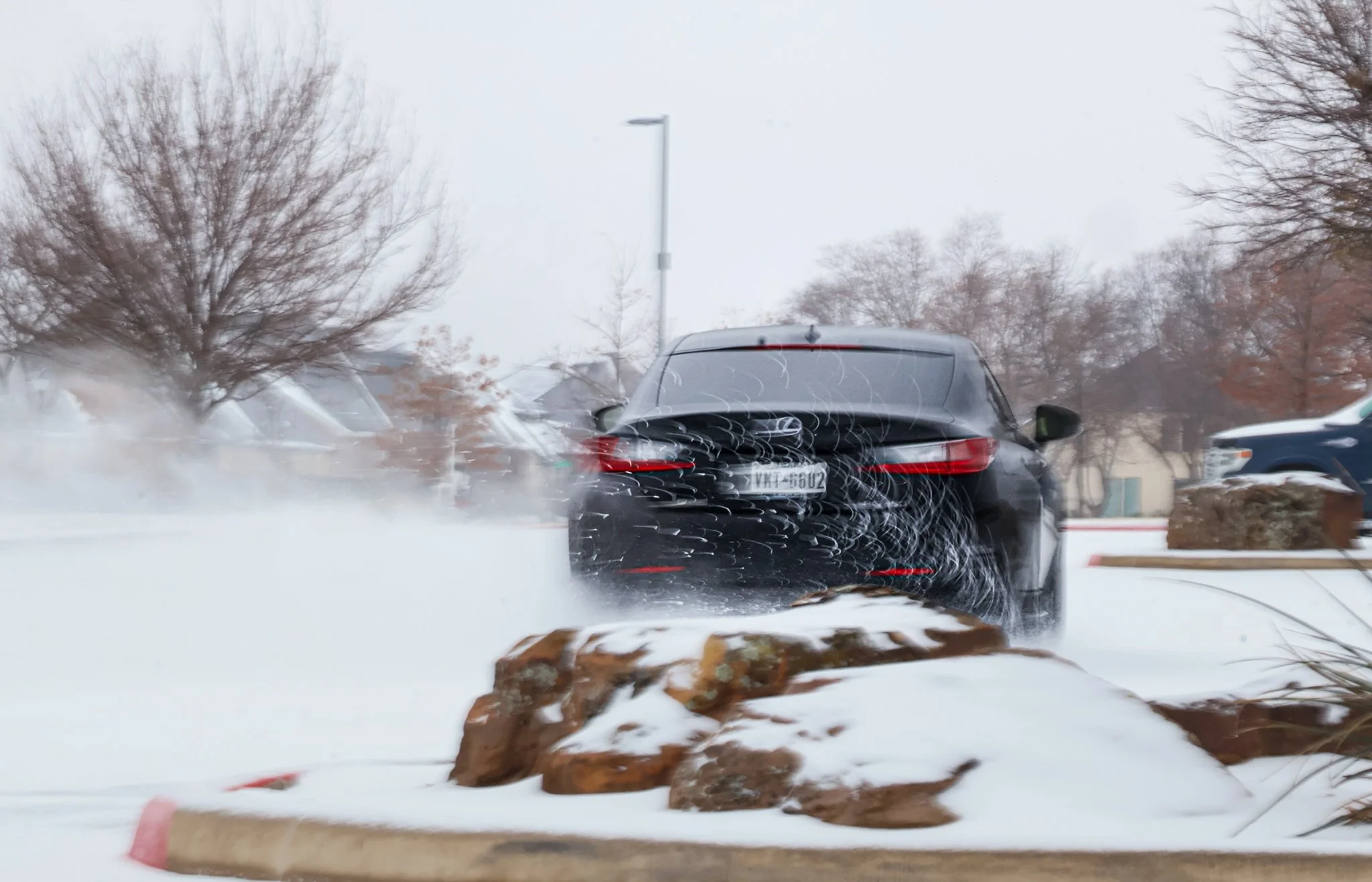 Black SUV driving through snowy parking lot, snow splashing behind it, leafless trees in the background.