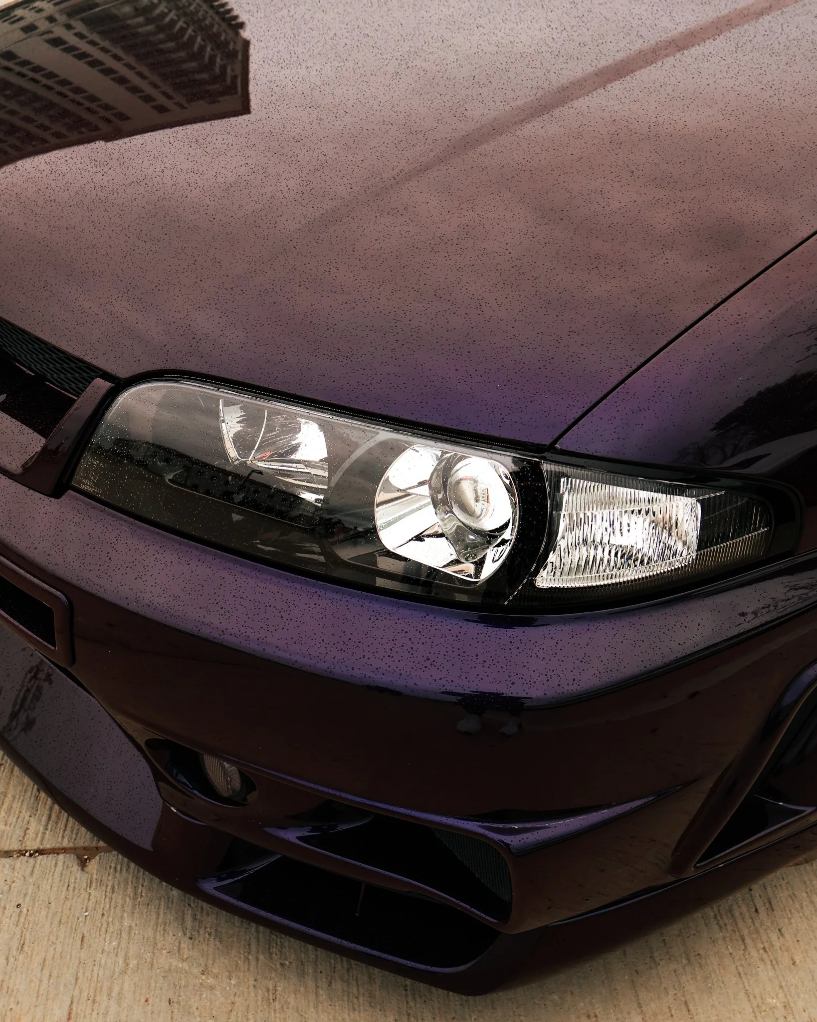 Close-up of a dark-colored car's front end, showing its headlight, part of the bumper, and hood with raindrops.