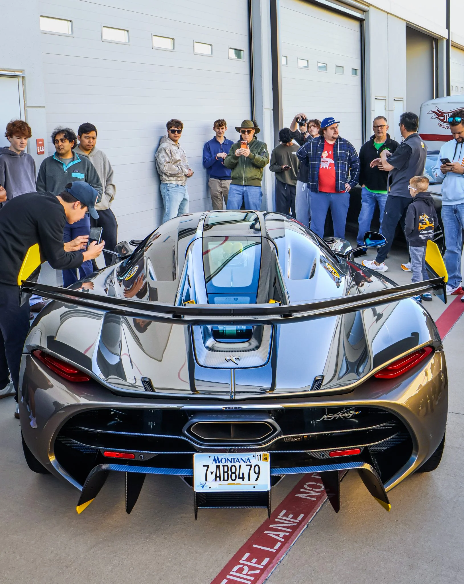 A sleek, black sports car with a Montana license plate, surrounded by a crowd of people taking photos and looking at the car in front of a garage.