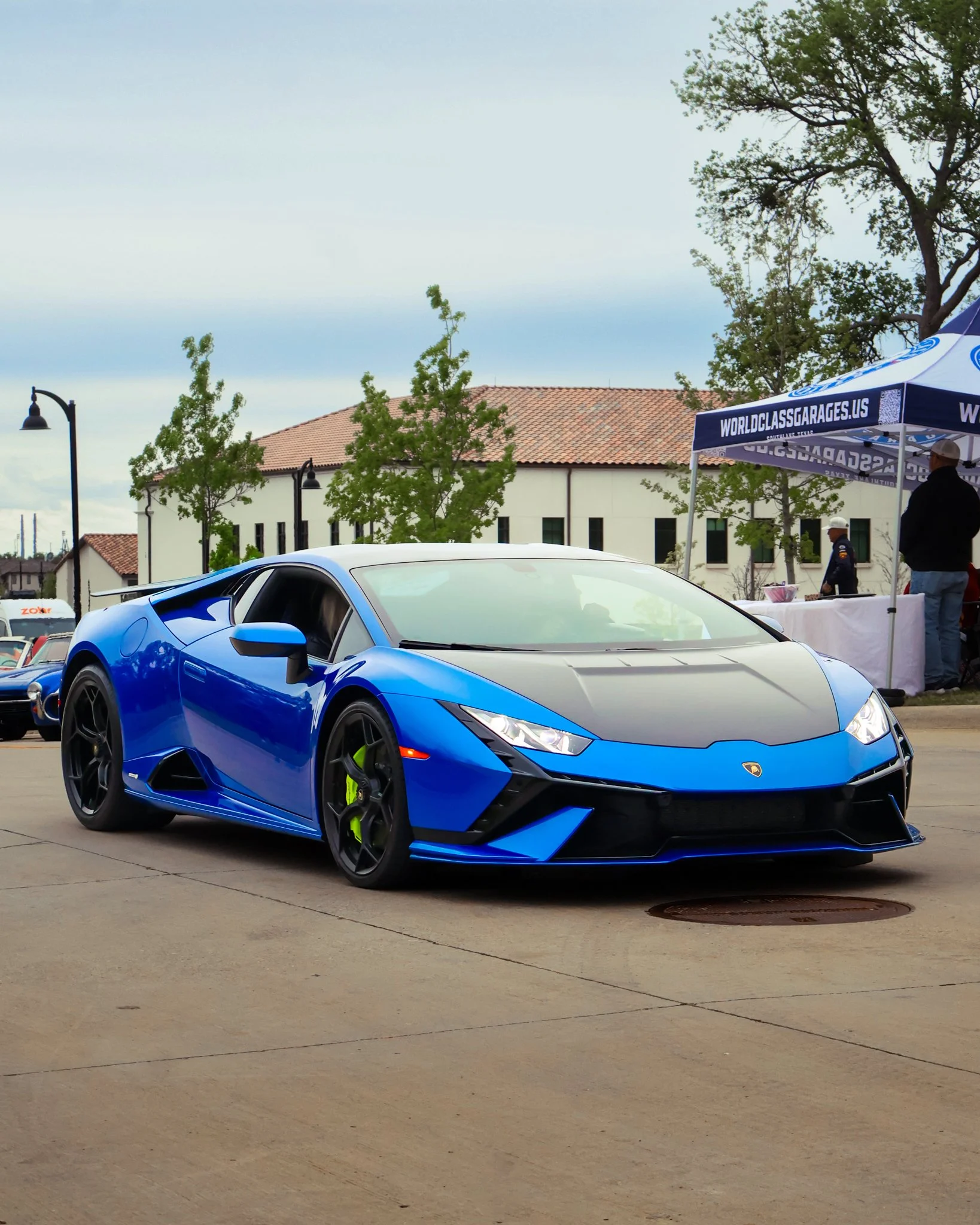 A blue Lamborghini sports car parked on a street during a car show, with a tent and people in the background.