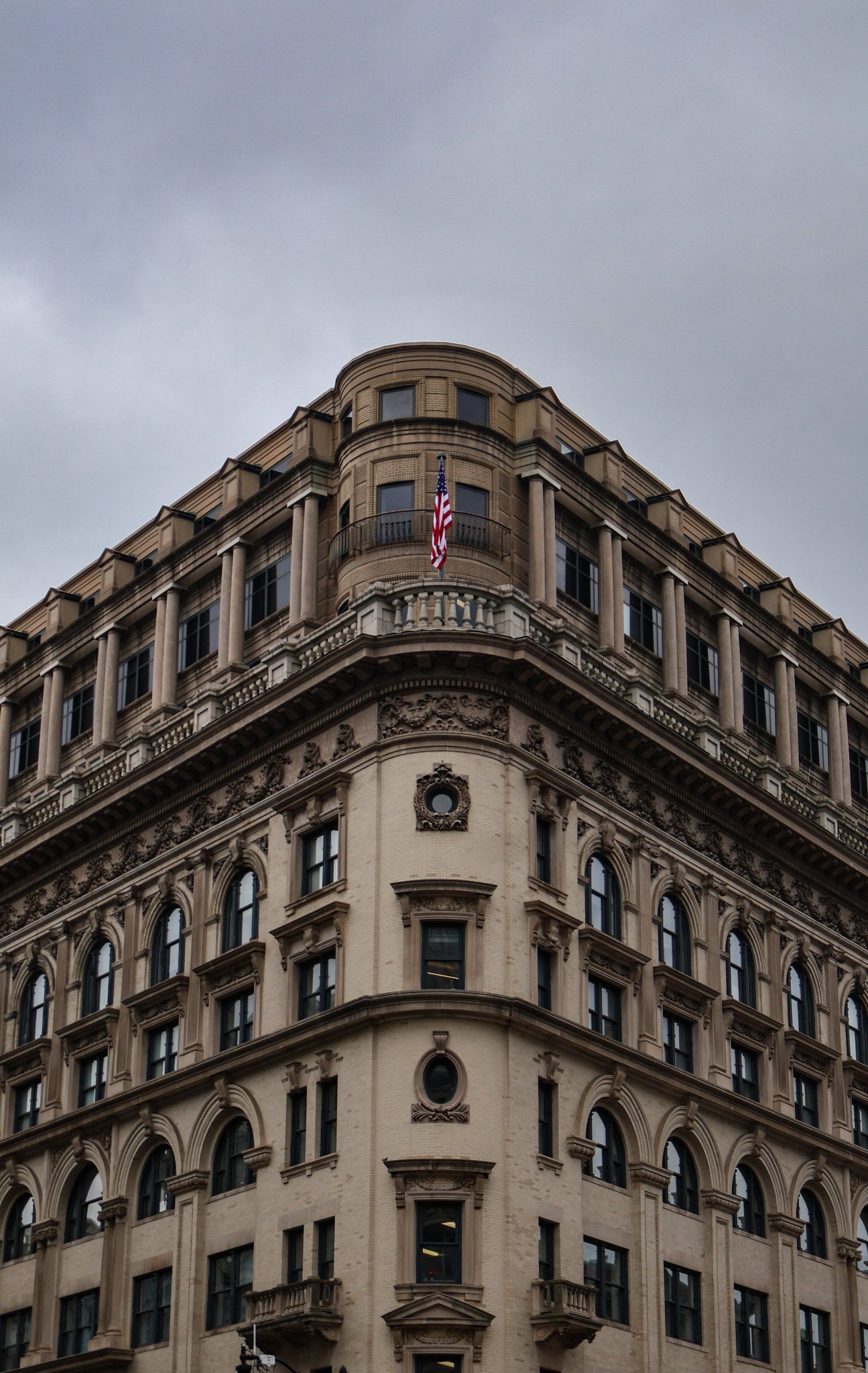 A historic multi-story building with ornate architectural details, a rounded corner, and an American flag flying on a small balcony, under a cloudy sky.
