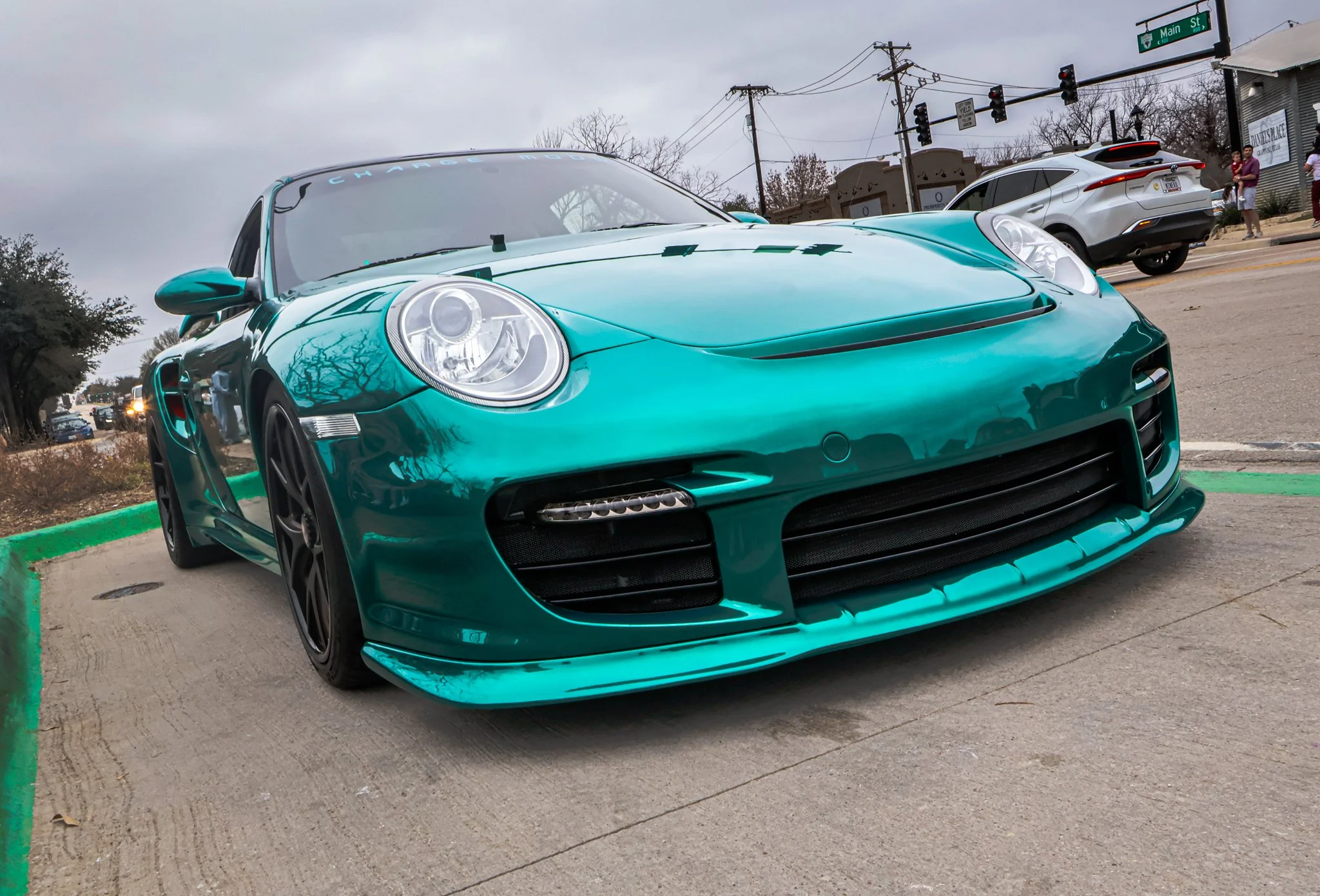 Teal Porsche sports car parked on the street, with a traffic light and other vehicles in the background.